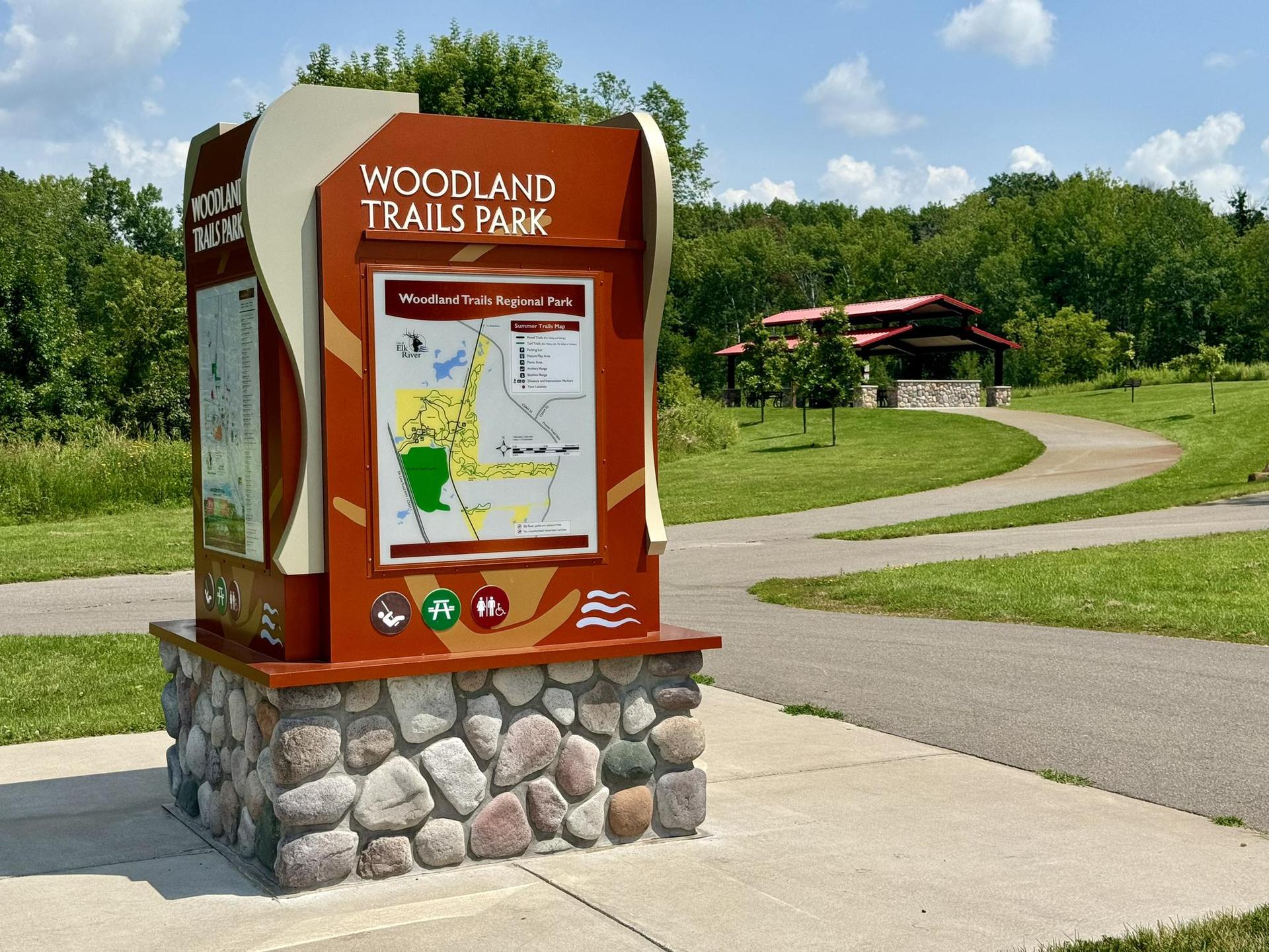 Information kiosk next to a paved path, with a building and trees in sunlight.
