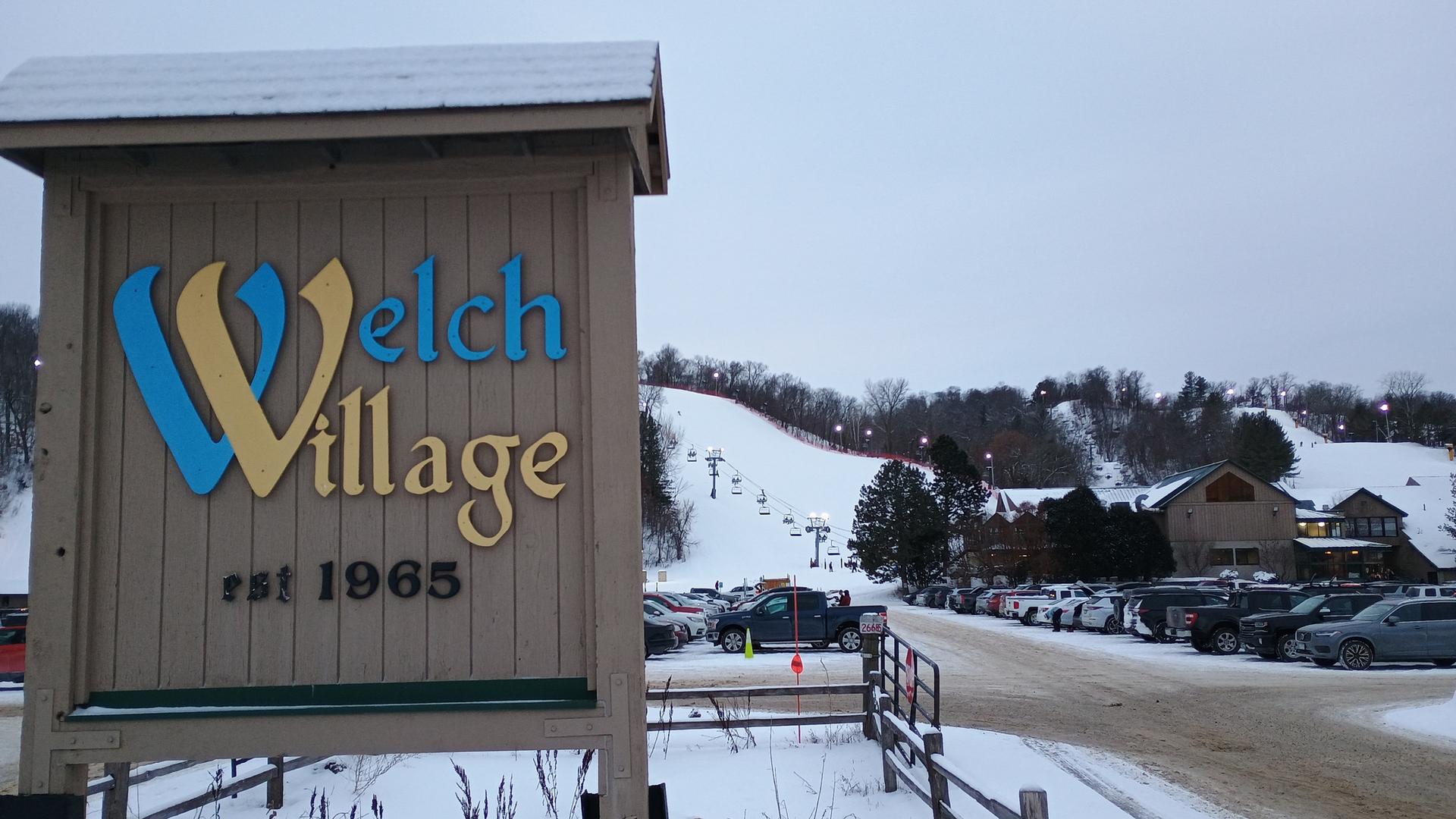 A ski resort sign in winter with snow-covered slopes and parked cars.