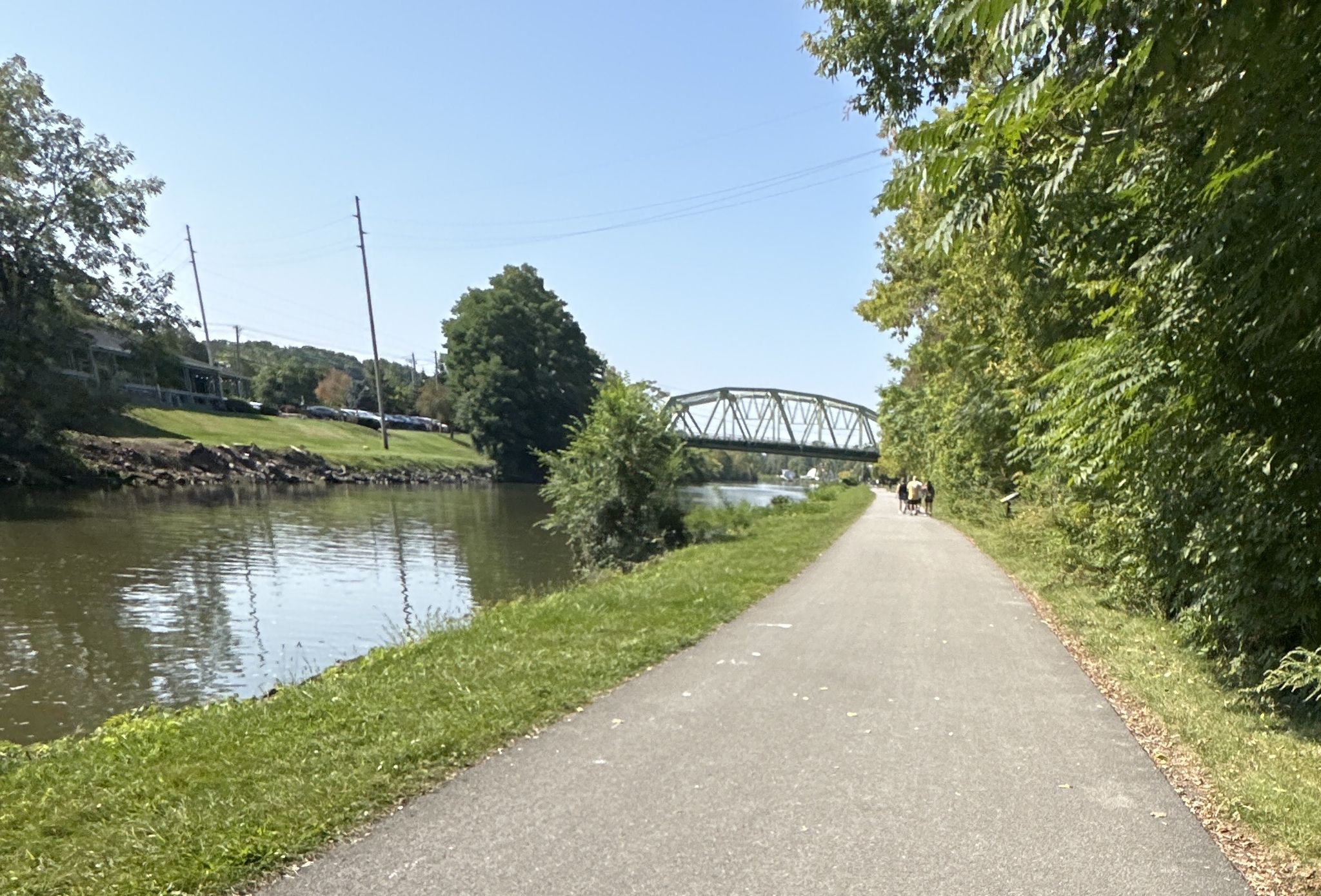 Paved path beside a river, leading to a green truss bridge with people. - Erie Canalway and Genesee Riverway Trail Loop