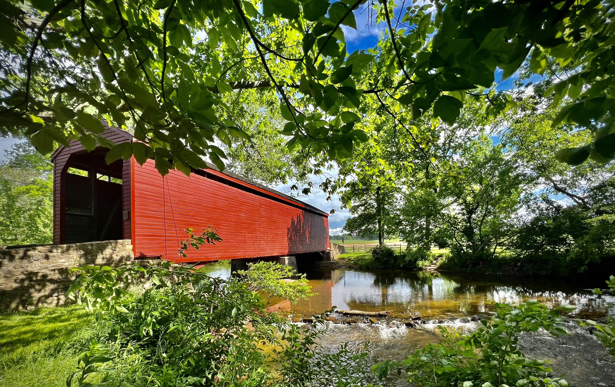 Red covered bridge spanning a stream, framed by lush green foliage. - North County Bridge Sampler
