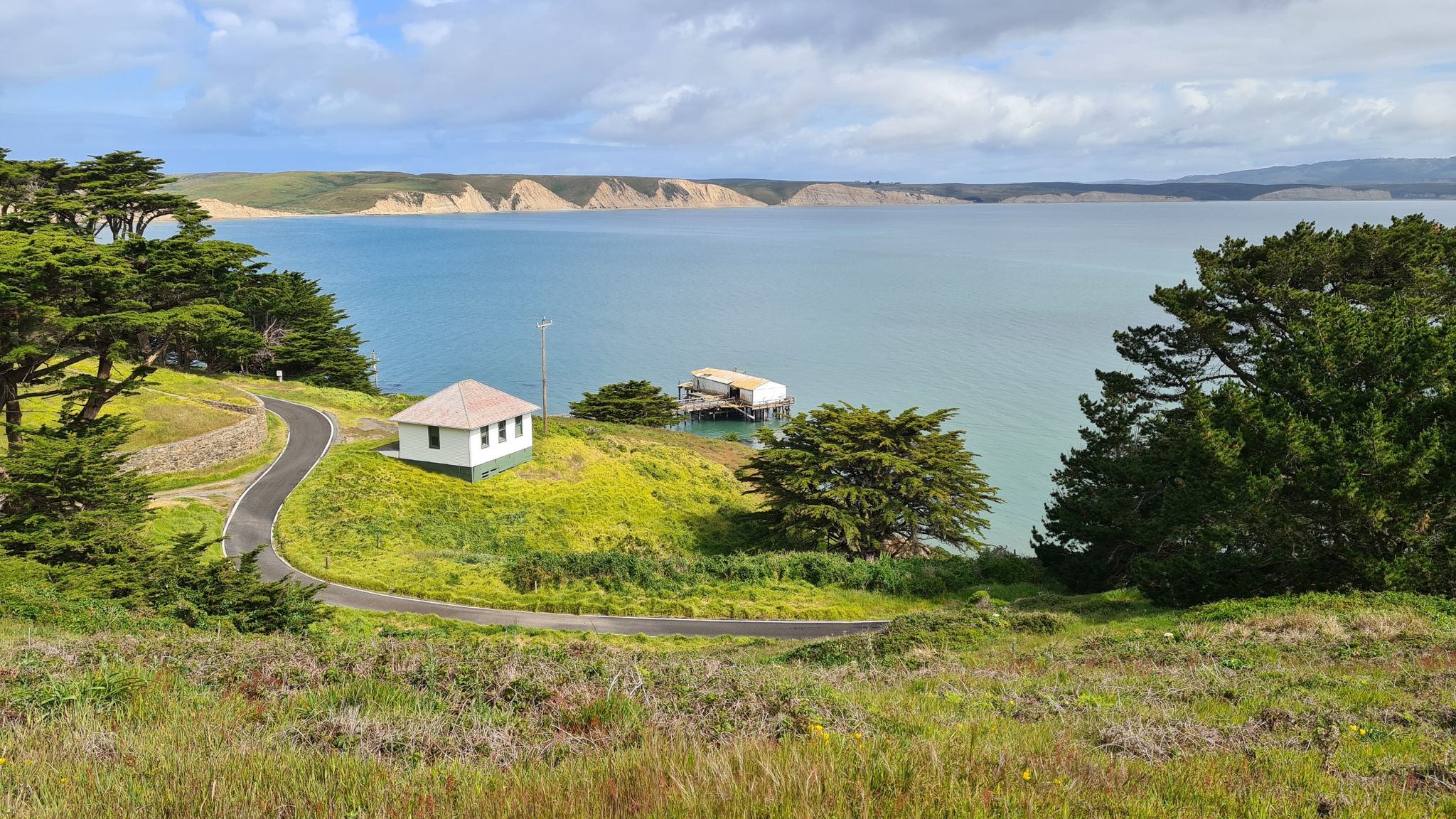 Coastal landscape with a bay, green hills, trees, and two small buildings. Bright, cloudy sky. - Chimney Rock Trail