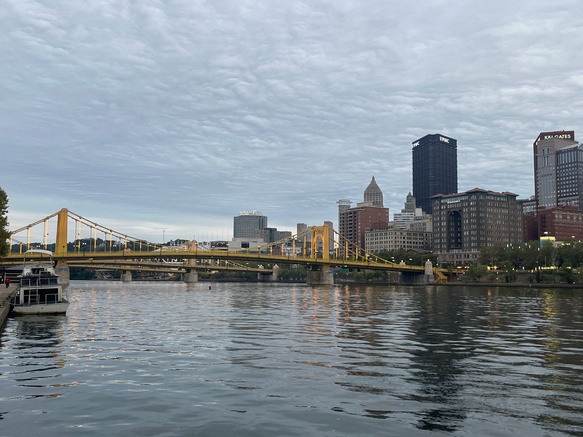 City skyline and yellow bridges over a river with a boat under cloudy skies. - Three Rivers Heritage Trail