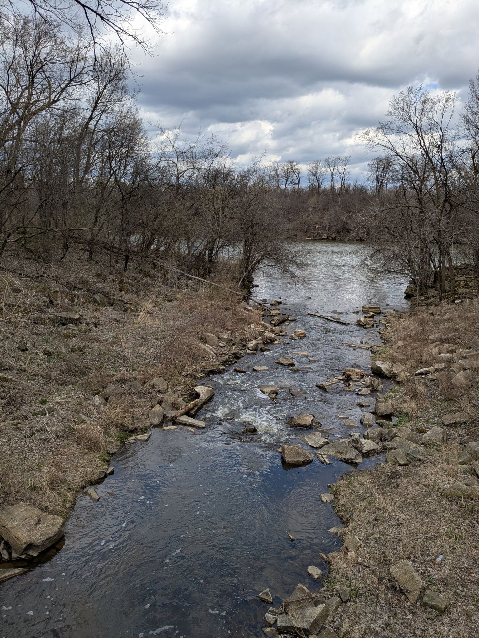 A rocky stream flowing into a wider river, surrounded by bare trees under a cloudy sky. - Cal-Sag Trail – Cal-Sag Channel loop from Site A