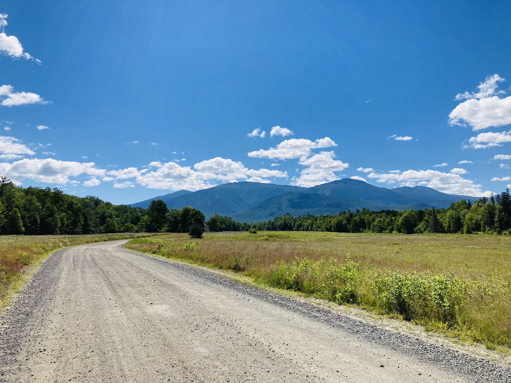 A wide gravel road curves through green fields towards distant mountains. - Old Cherry Mountain Road, Valley Road, & Jefferson Notch Road gravel loop