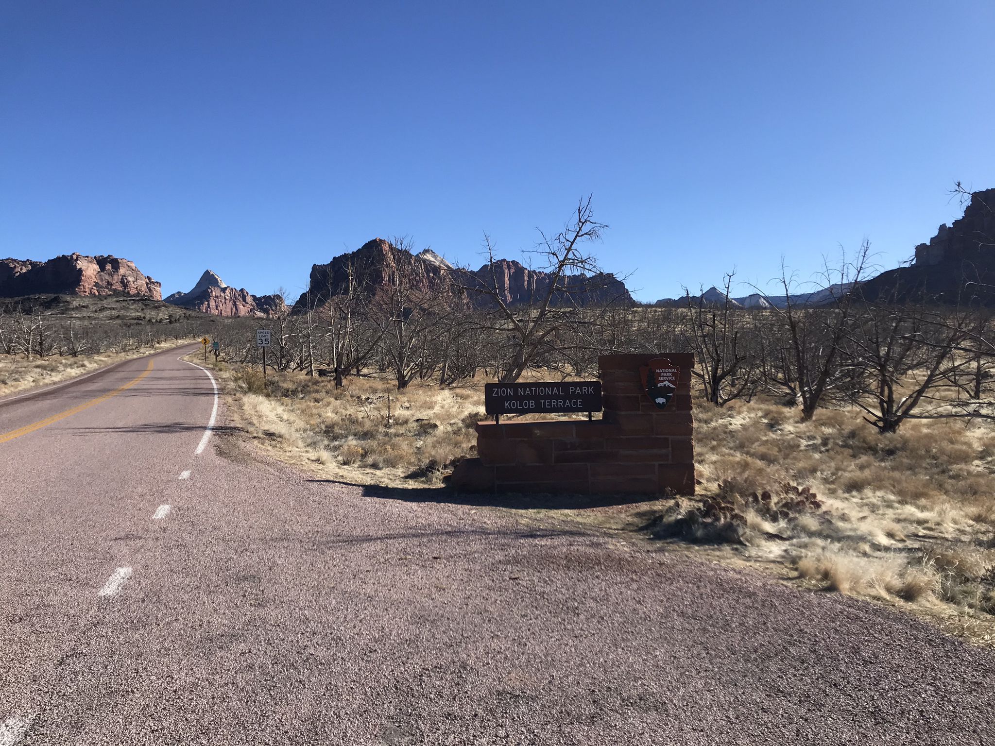 Road approaching a park entrance sign with red rock mountains beyond. - Kolob Terrace Road – Tabernacle Dome & Lambs Knoll loop from Virgin