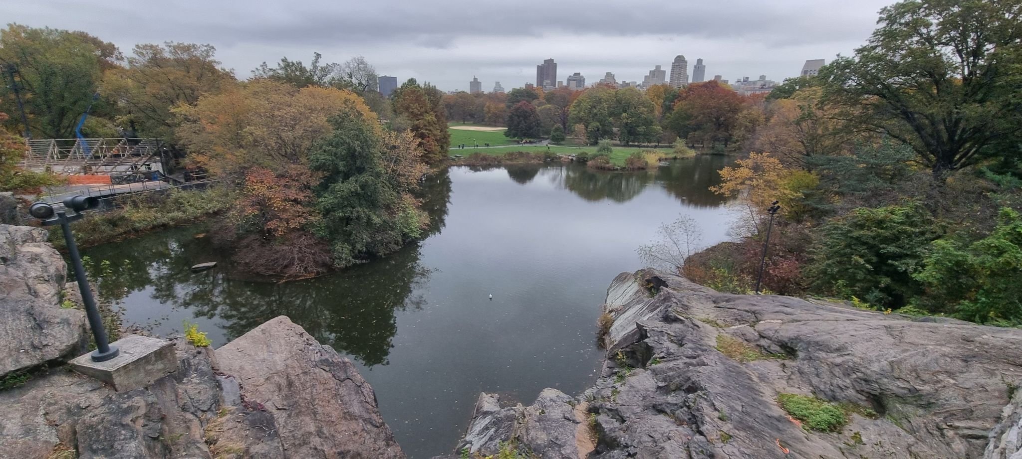 Lake viewed from rocky outcrop, autumn trees, distant city, cloudy day. - Central Park Lake and Bow Bridge Loop