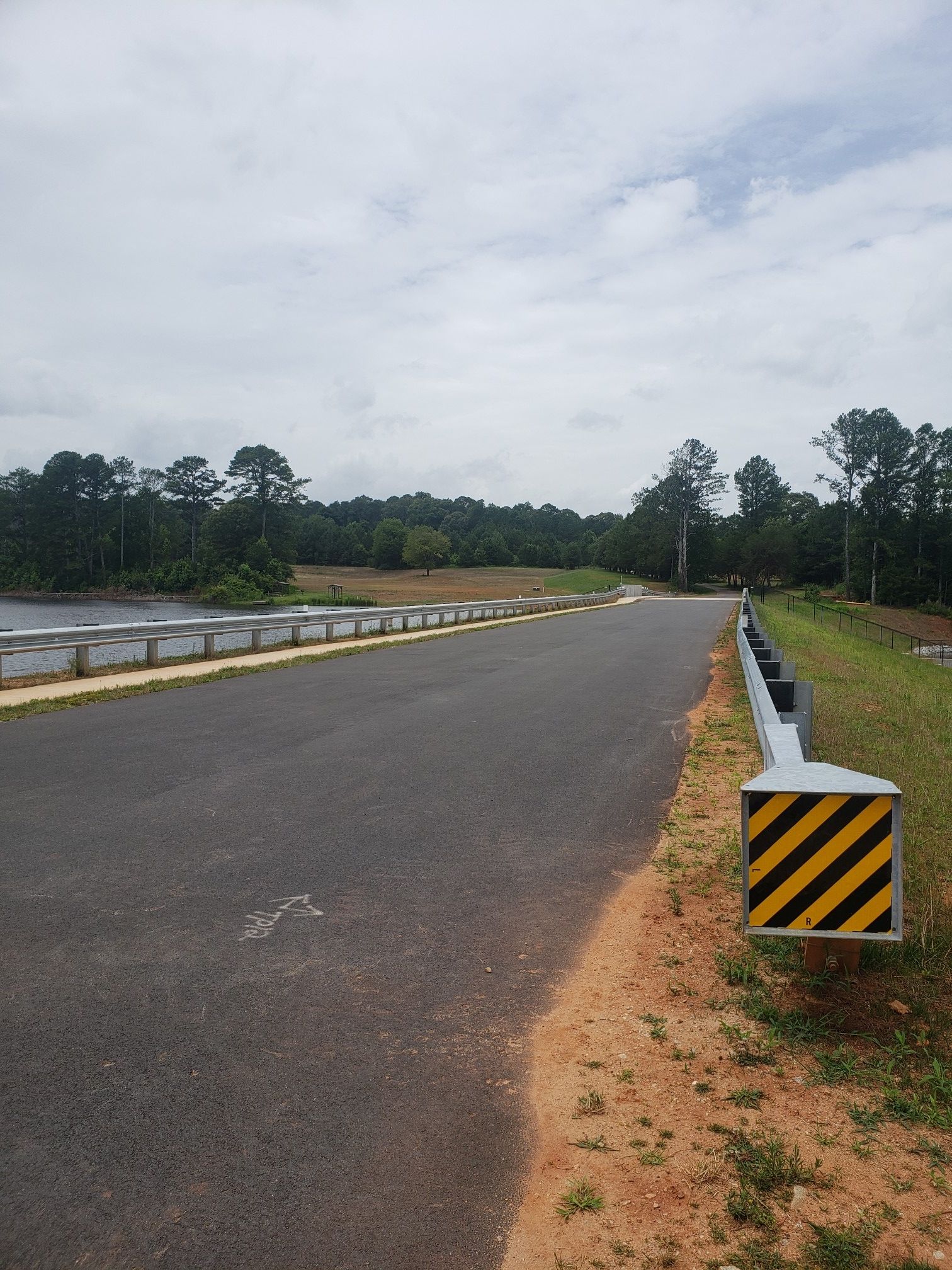 Paved road with guardrails, striped barrier, and forested landscape under cloudy sky. - Panola Mountain Greenway