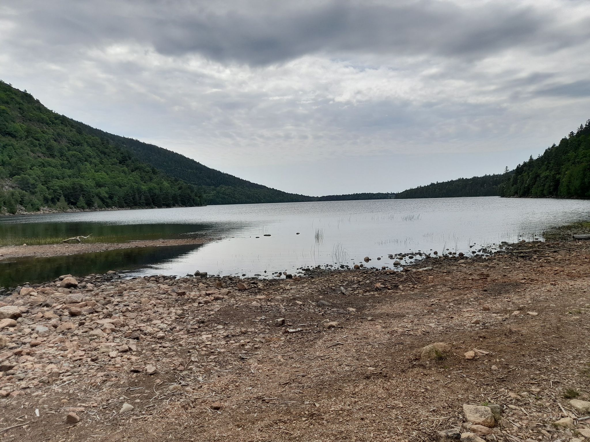 Overcast day, calm lake with a rocky shore, surrounded by forested hills. - Jordan Pond Path and South Bubble Trail