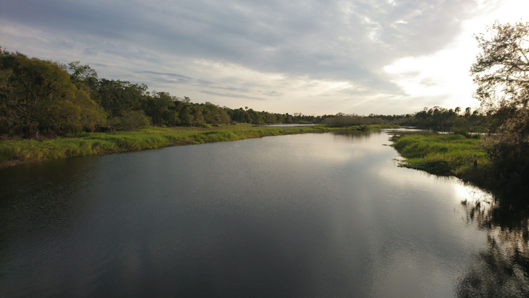 A wide, calm river flows between green banks under a bright, cloudy sky. - Palmetto Ridge Campground Paved Route