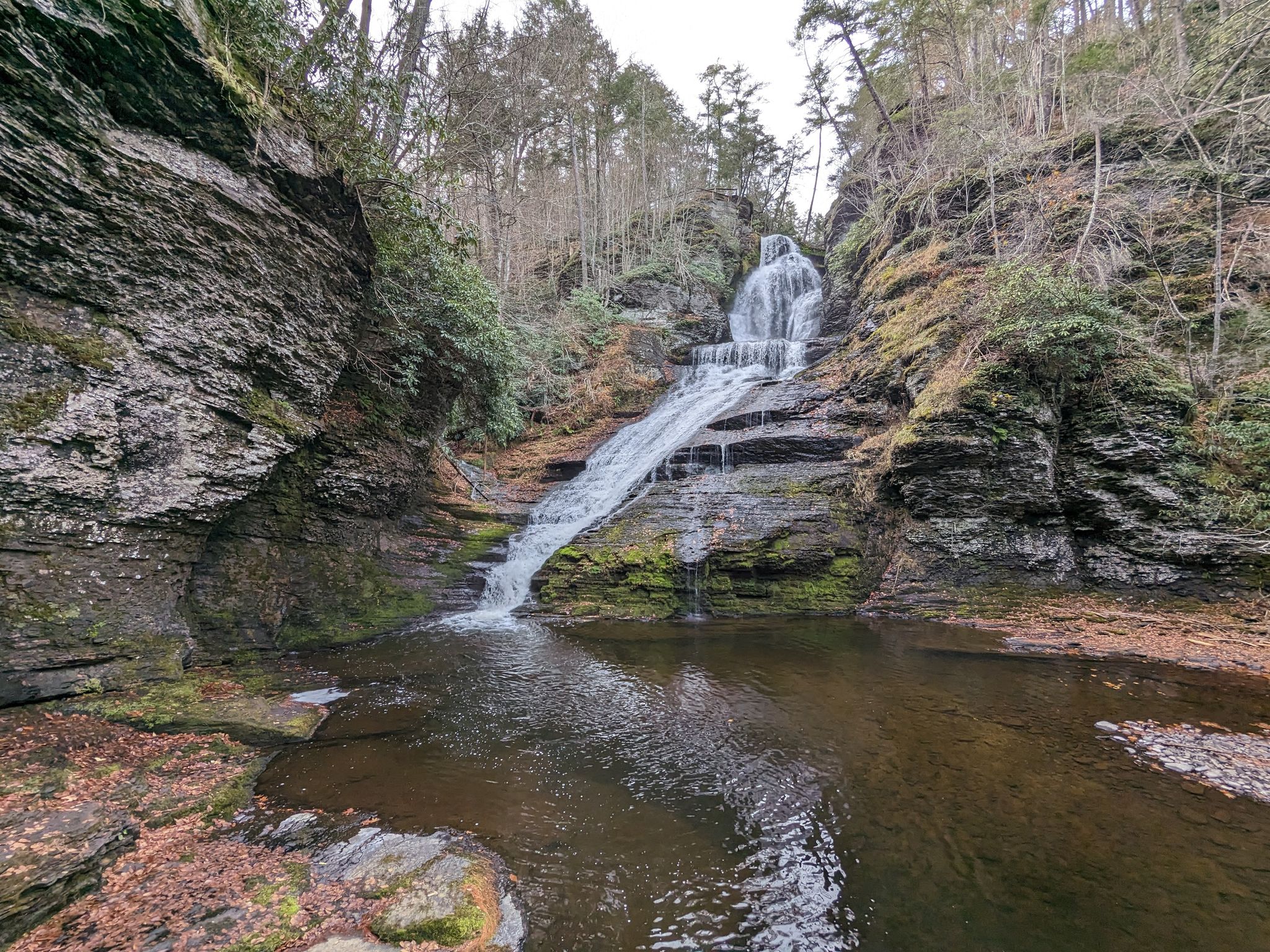 Waterfall cascades over rocky cliffs into a dark pool under an overcast sky. - Dingmans Creek Trail