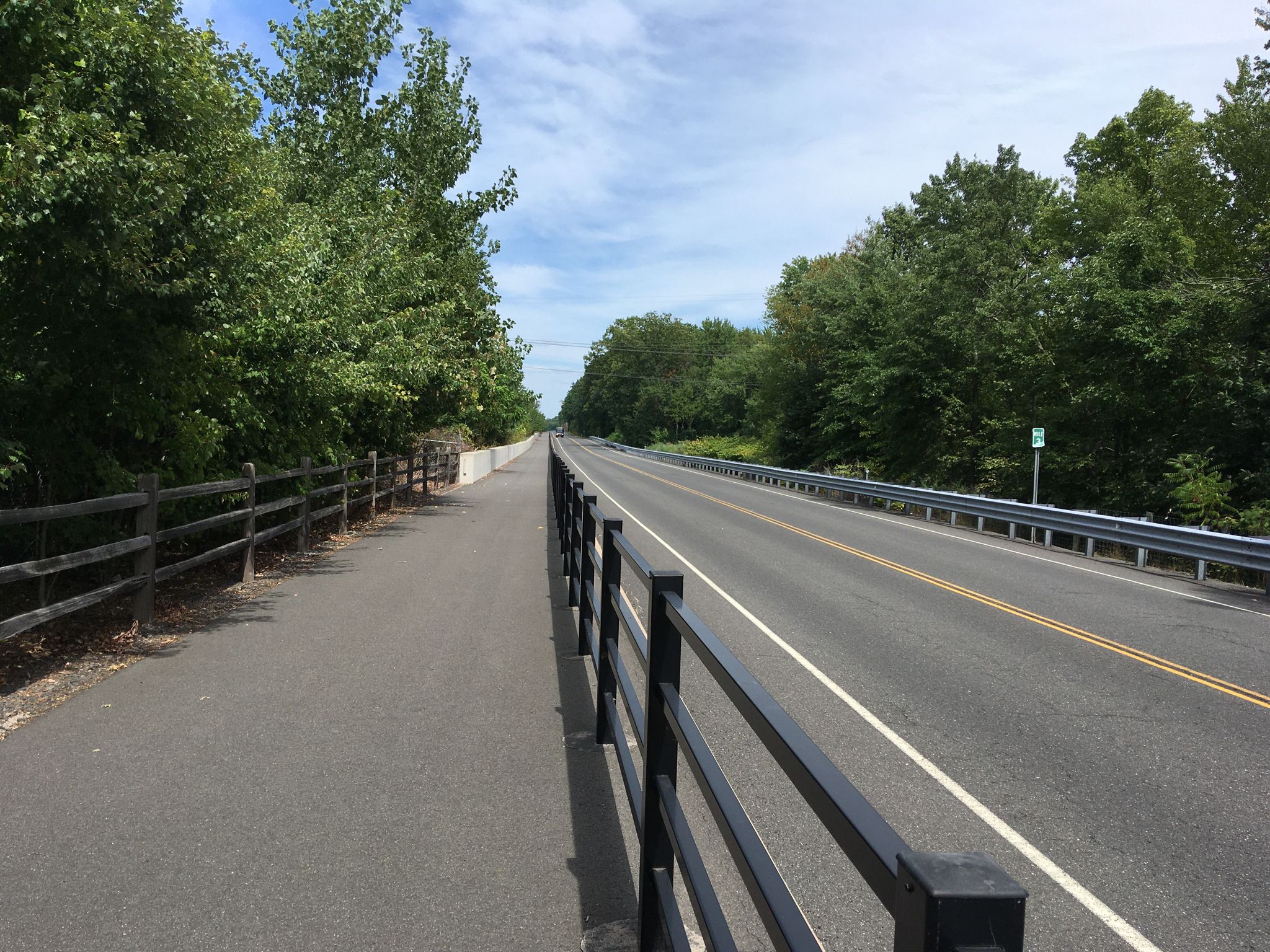 Paved path and road separated by fences, lined with green trees under a bright sky. - CTfastrak Multi-Use Trail