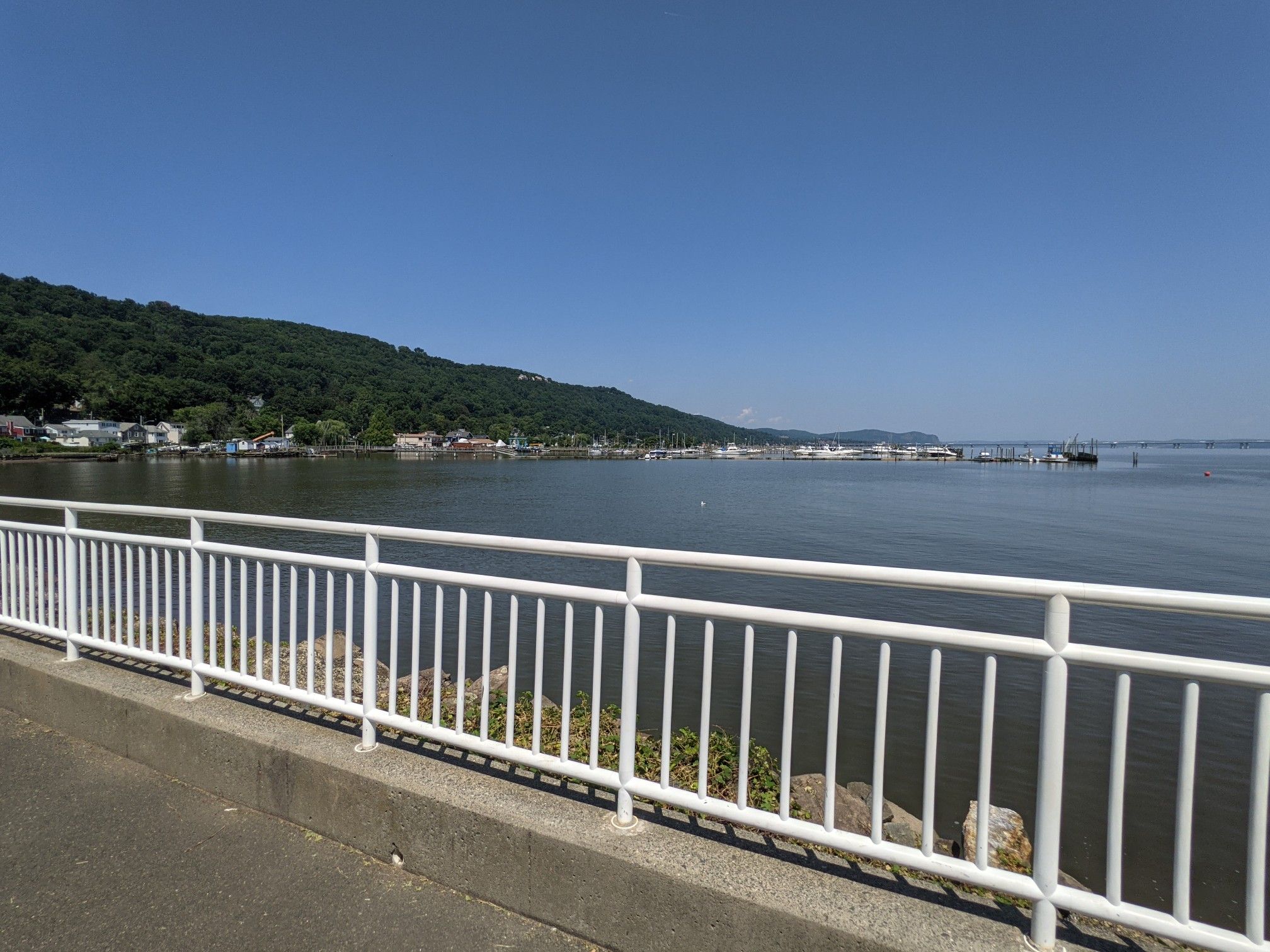 Sunny view of a marina and forested hills across calm water, with a white railing. - Piermont Pier