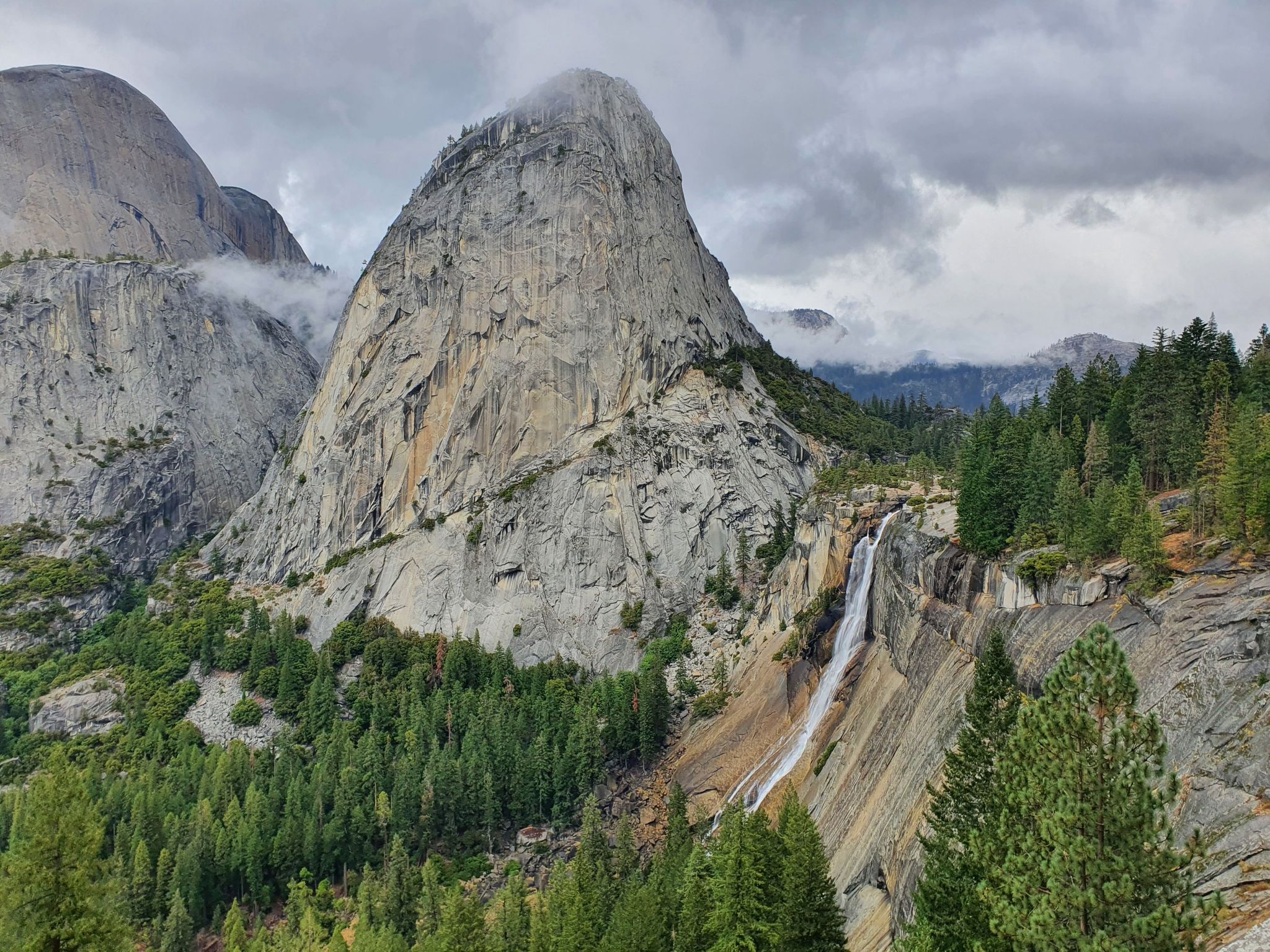Large granite mountain and waterfall surrounded by pine forest under an overcast sky. - Vernal and Nevada Falls via Mist Trail