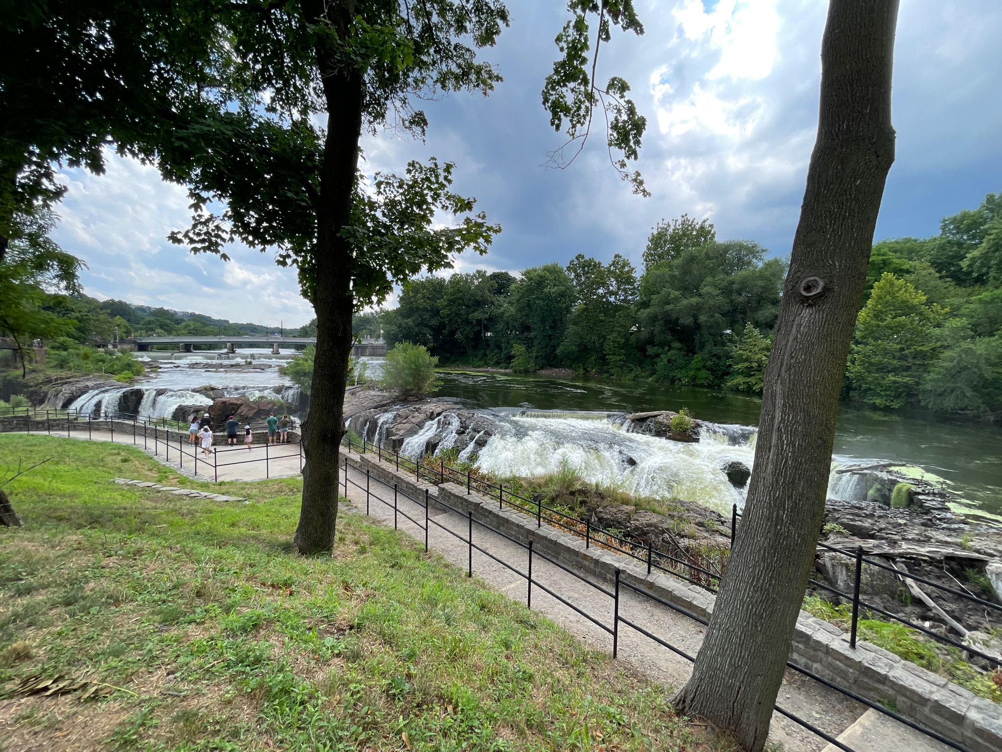 People view multiple waterfalls from a paved path, surrounded by green trees. - Paterson Falls Park Trails