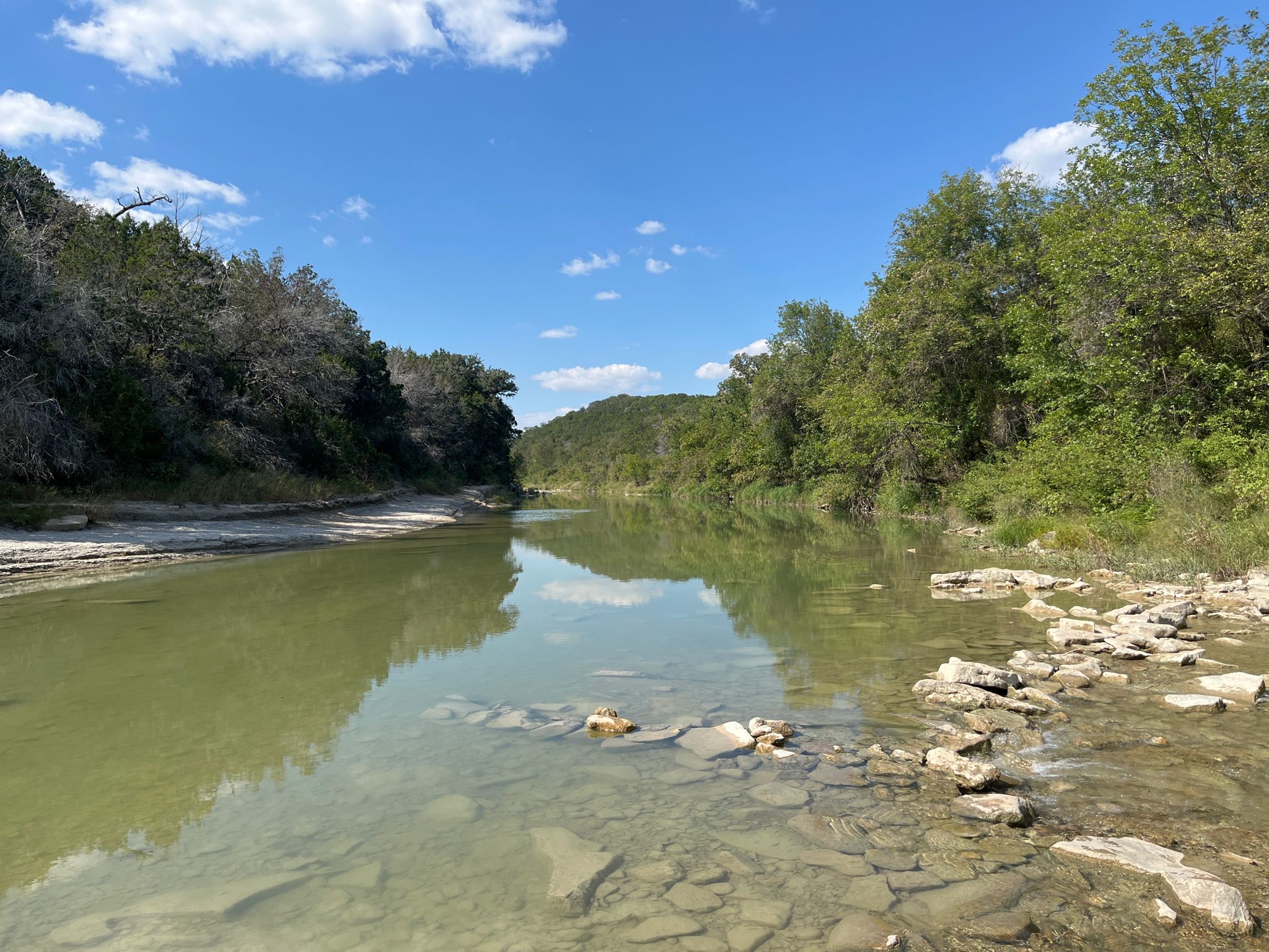 A calm river with rocky shores reflects the sky, surrounded by green hills. - Dinosaur Valley State Park – Eroded Trail Section loop from Dinosaur Valley State Park