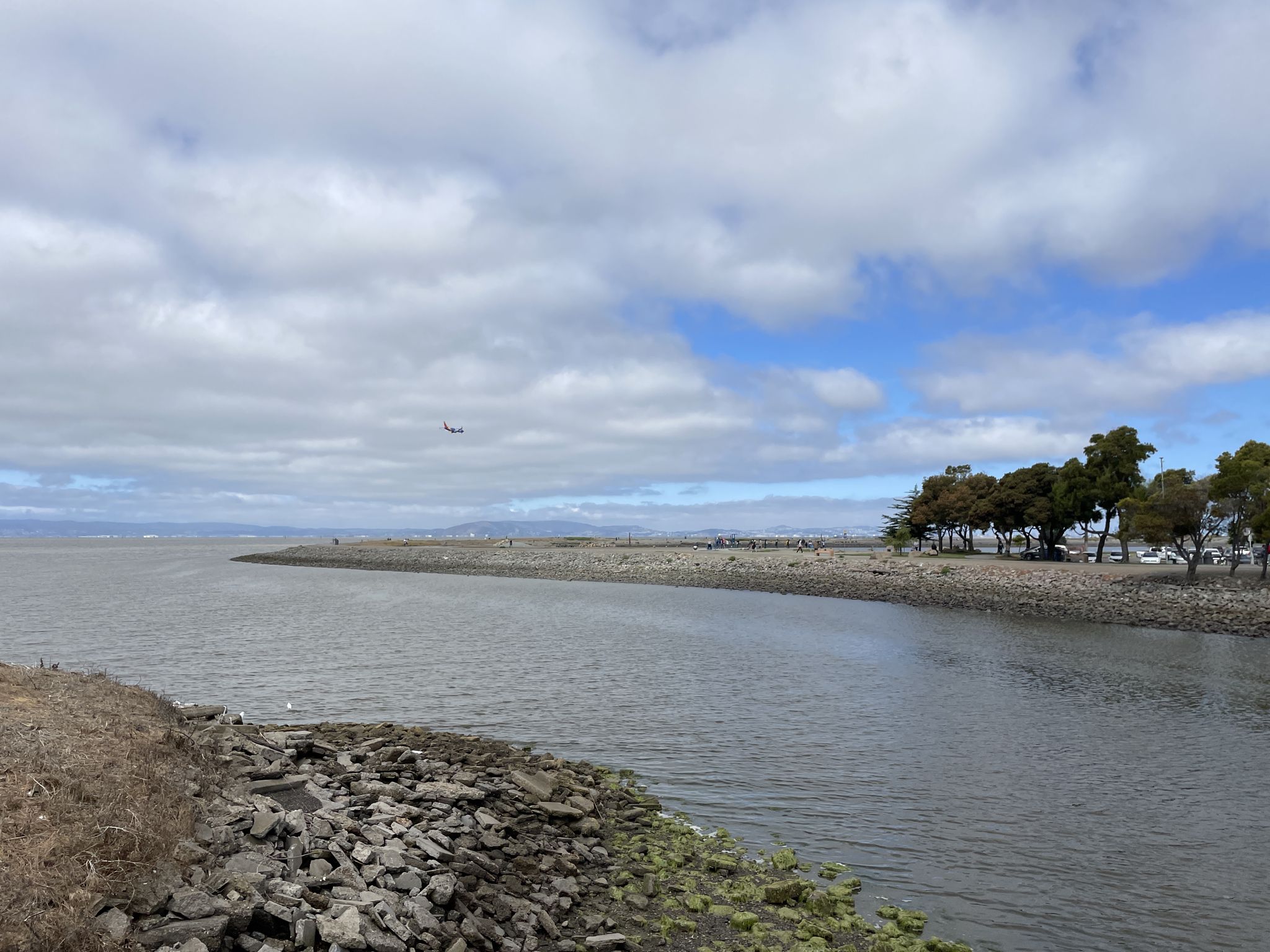 A wide bay with a rocky shore under a cloudy sky, an airplane overhead. - Heron Bay – San Francisco Bay Trail loop from Arroyo High School