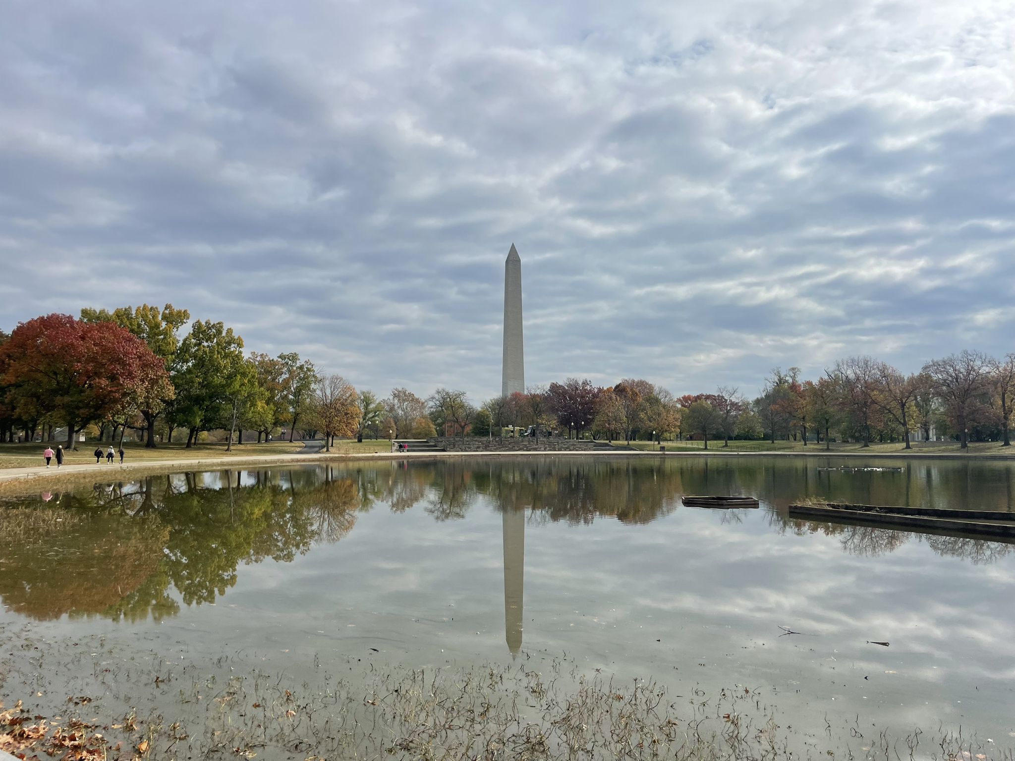 Obelisk reflecting in water, autumn trees, cloudy sky, distant people. - White House – Washington Monument loop from Arlington