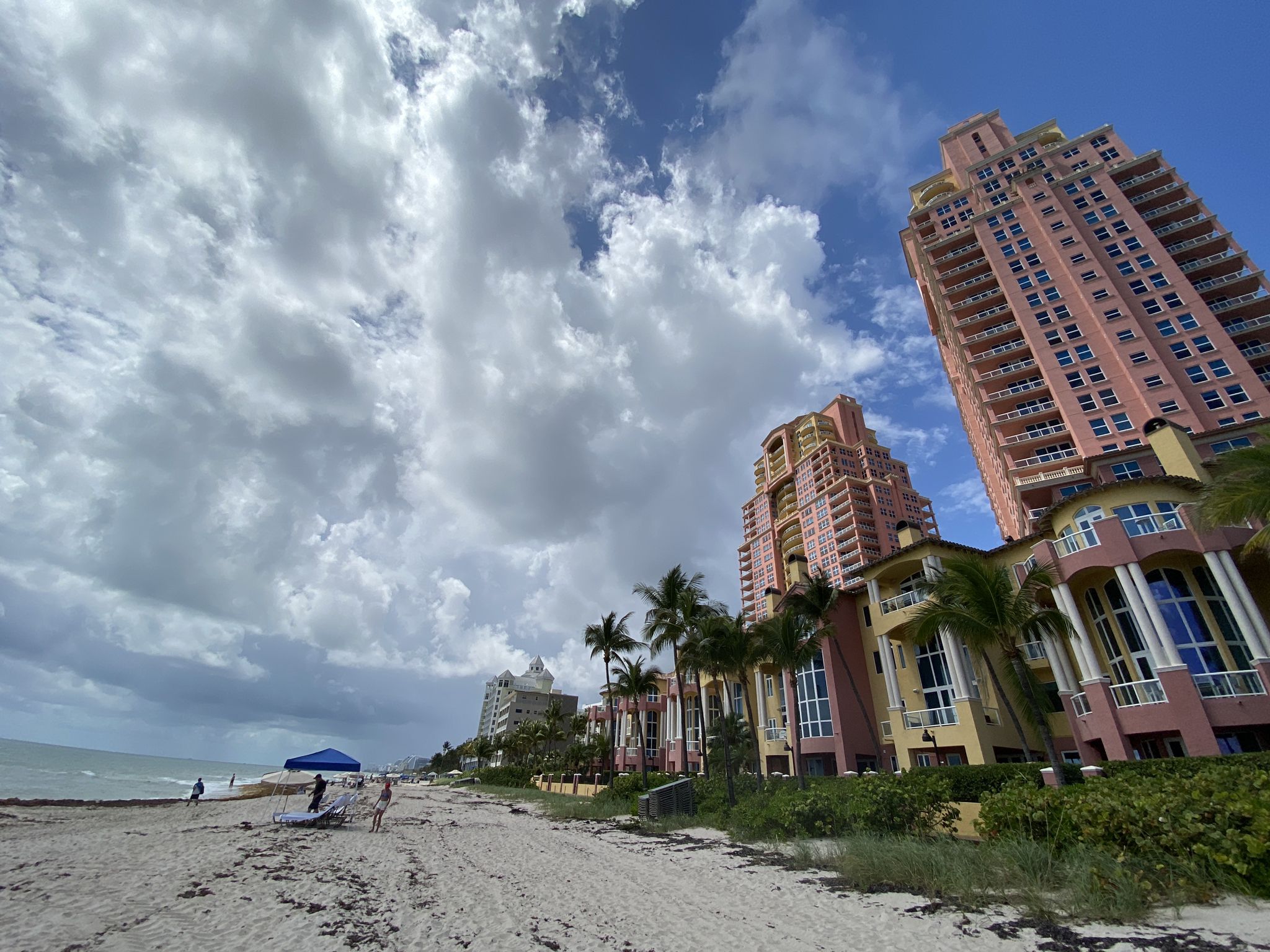 Beach with tall, colorful buildings, palm trees, and people under a cloudy sky. - Barrier Islands Greenway