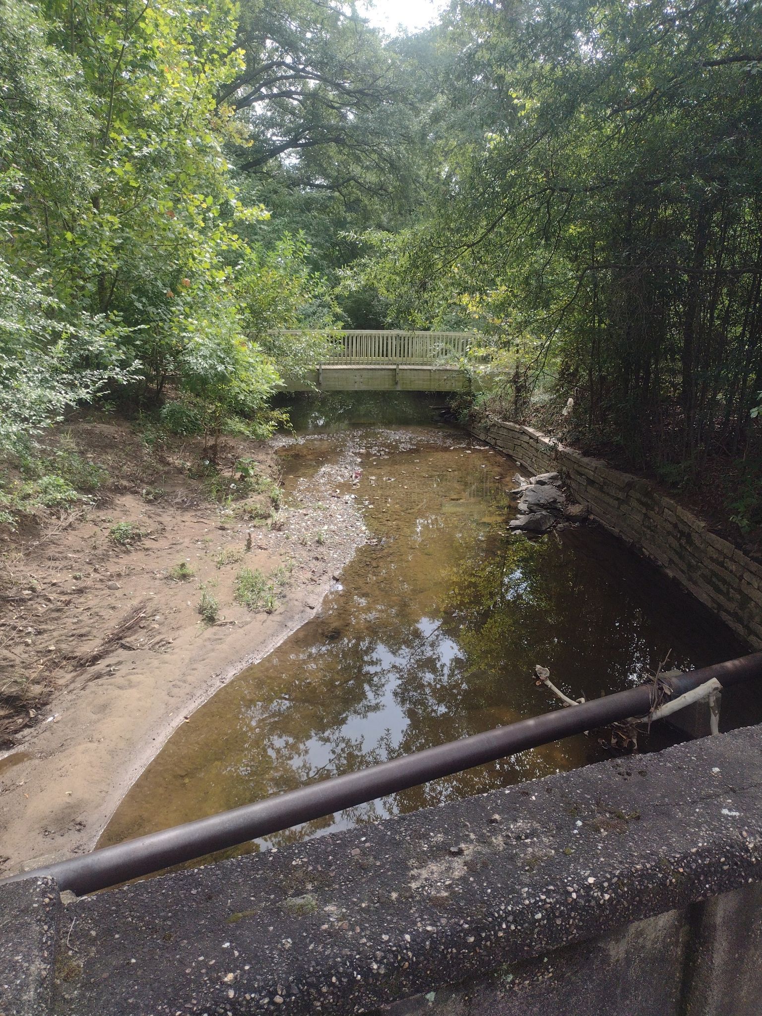 A small stream flows under a wooden bridge surrounded by green trees. - East Coast Greenway and American Tobacco Trail Loop