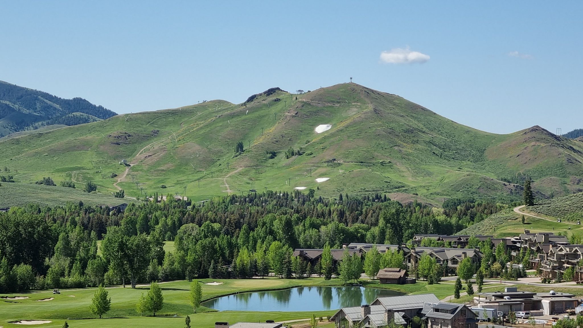 Sunny view of a green mountain, lake, golf course, and buildings. - Sun Valley Village loop from Sun Valley