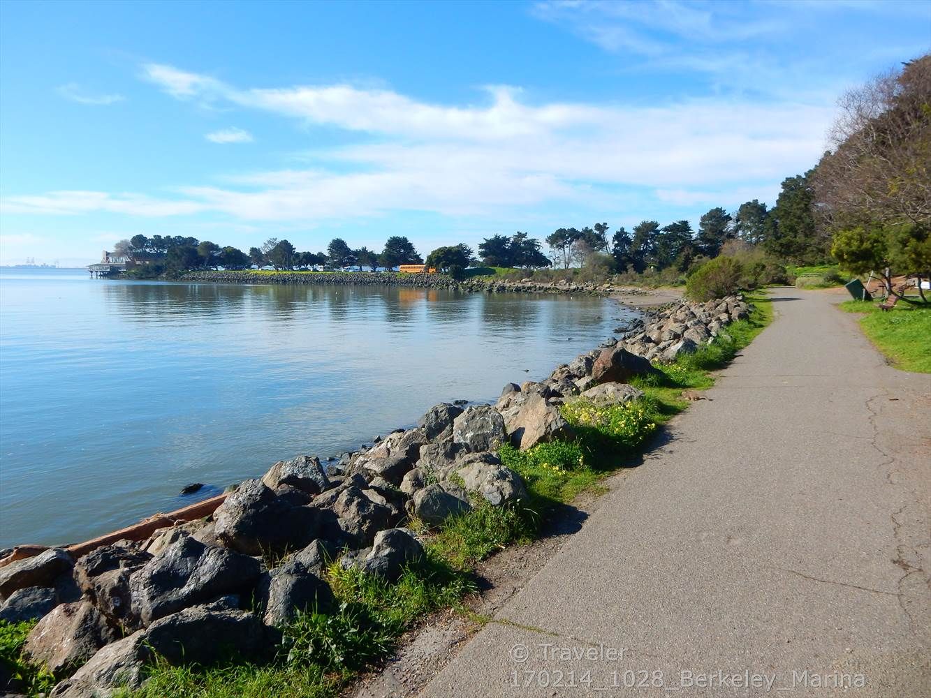 Paved path alongside rocky shore, calm water, and distant treeline under blue sky. - Aquatic Park – Berkeley Meadow loop from Emeryville
