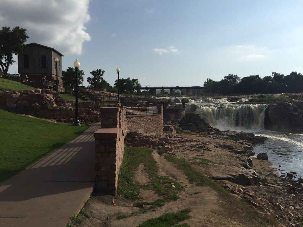 Sunny view of a waterfall, old building, and paved path beside a rocky river. - Falls Park Waterfalls – Sioux Falls Bike Trail loop from Augustana University