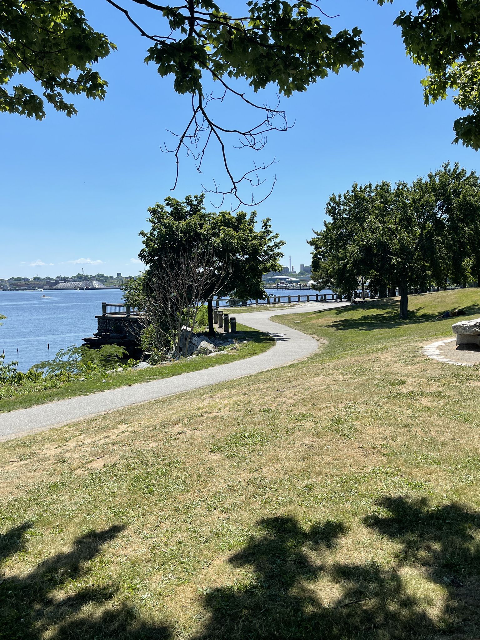 A paved path winds through a sunny park overlooking a calm water body. - Oakland Beach to India Point Park