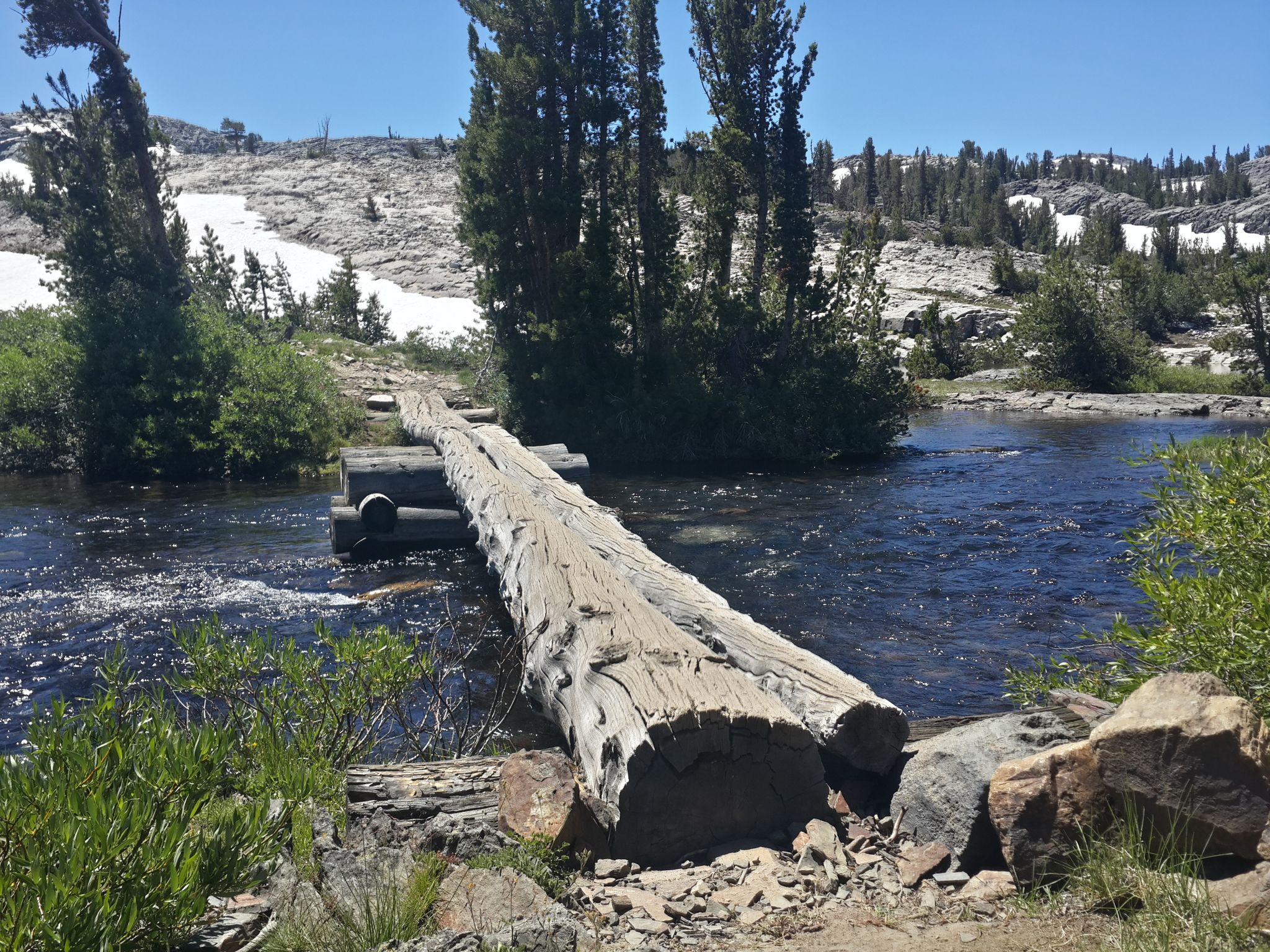 Rustic log bridge crosses a flowing river in a sunny, forested mountain landscape. - Shadow Creek Trail