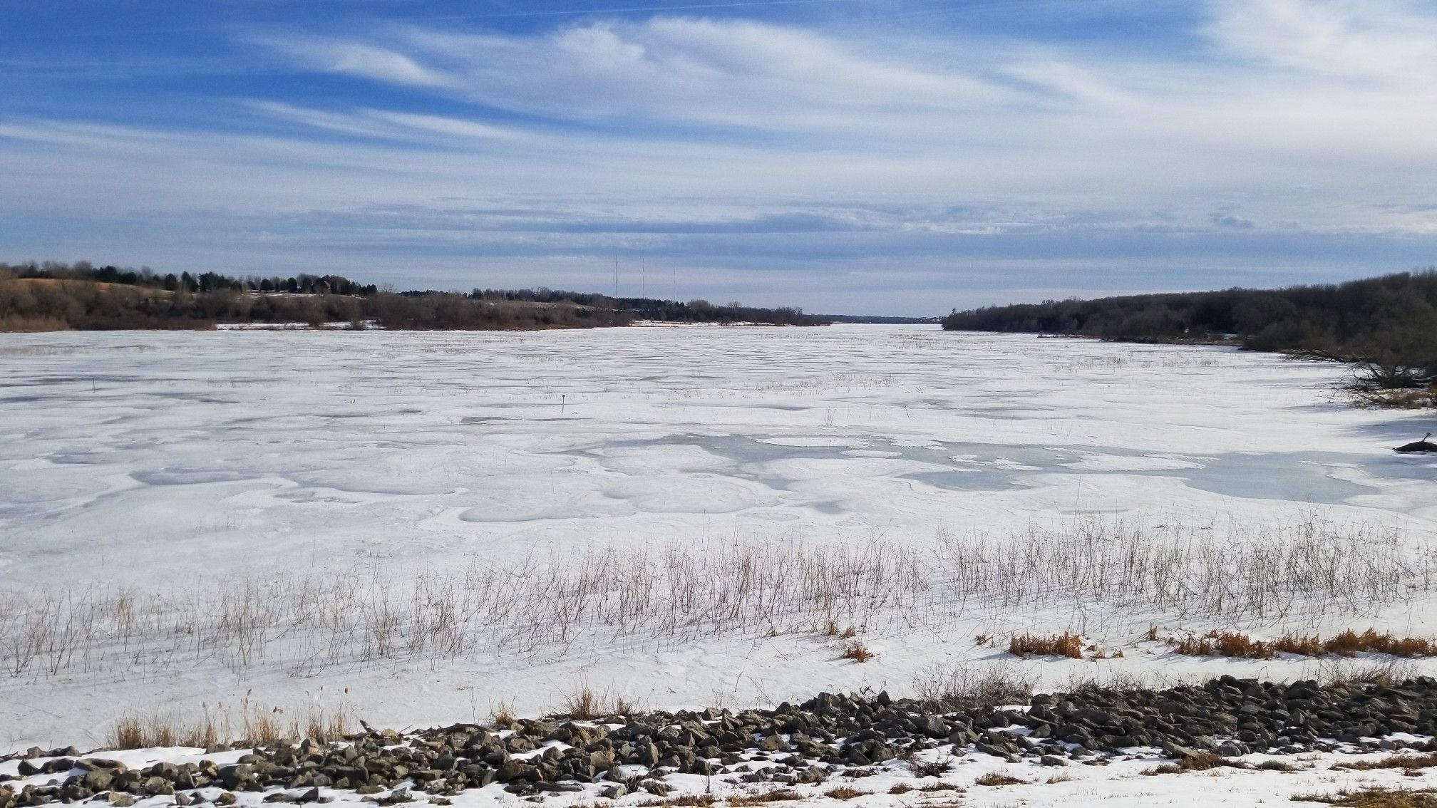 Expansive frozen lake with snow-covered banks and distant trees under a blue sky. - Glenn Cunningham Lake – Cunningham Lake Bridge loop from Papio Creek D-4 Dam