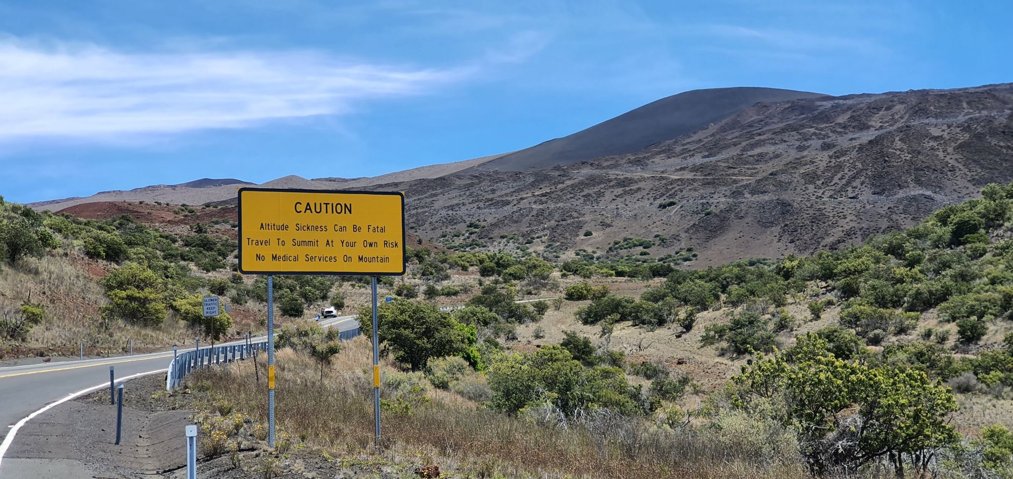 A yellow caution sign stands beside a road in a hilly, vegetated landscape. - Mauna Kea Access Road to Maunakea Visitor Information Station