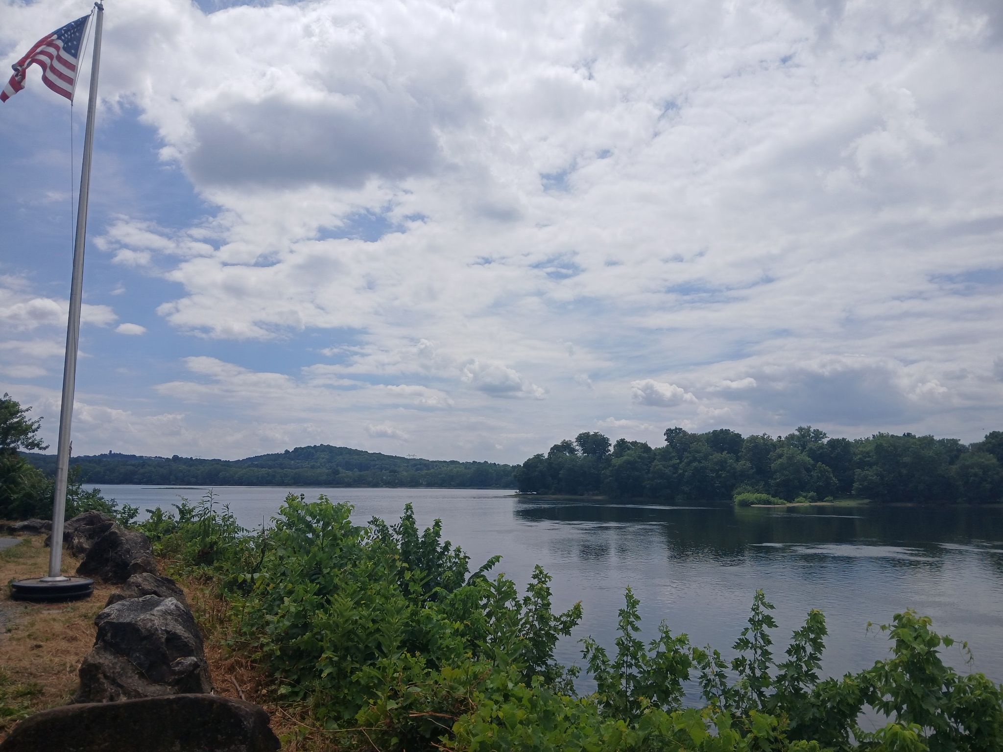 American flag overlooks a wide river with forested banks under a cloudy sky. - Northwest Lancaster County River Trail