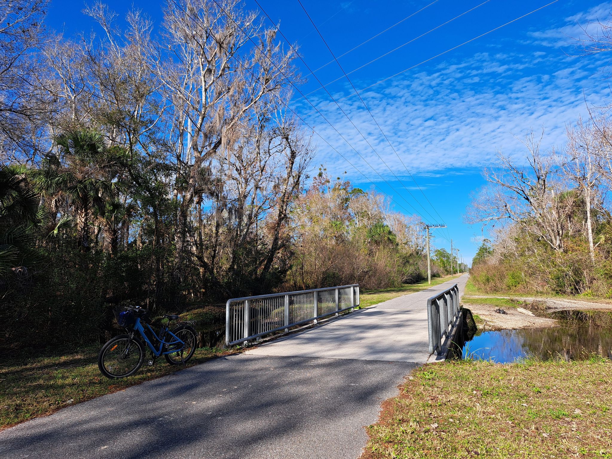 Paved path with a small bridge, bicycle parked, under a bright blue sky. - East Central Rail Trail loop from Mims