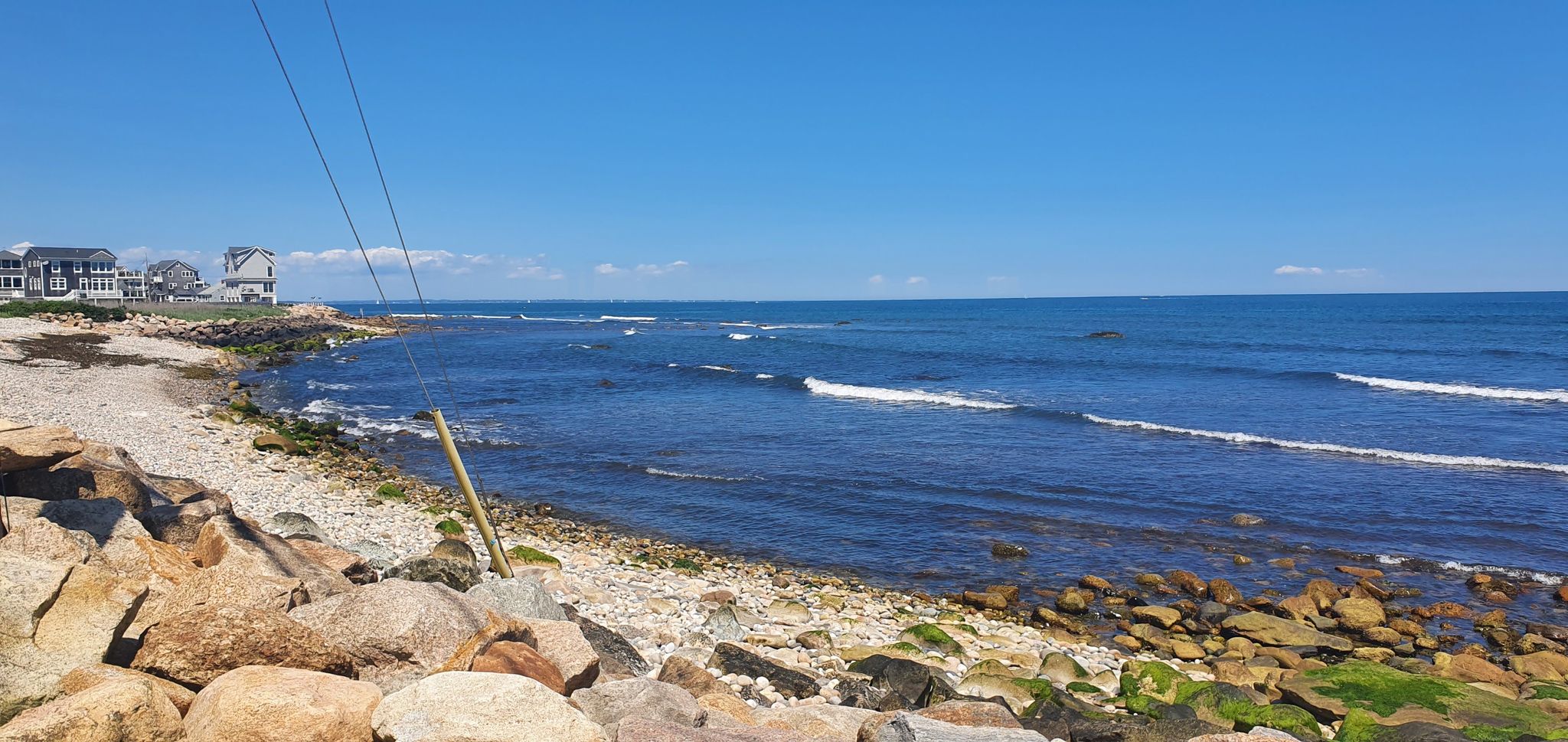 A sunny view of a rocky coastline with blue ocean and distant houses. - The Towers (Narragansett) – Point Judith Lighthouse loop from Saunderstown