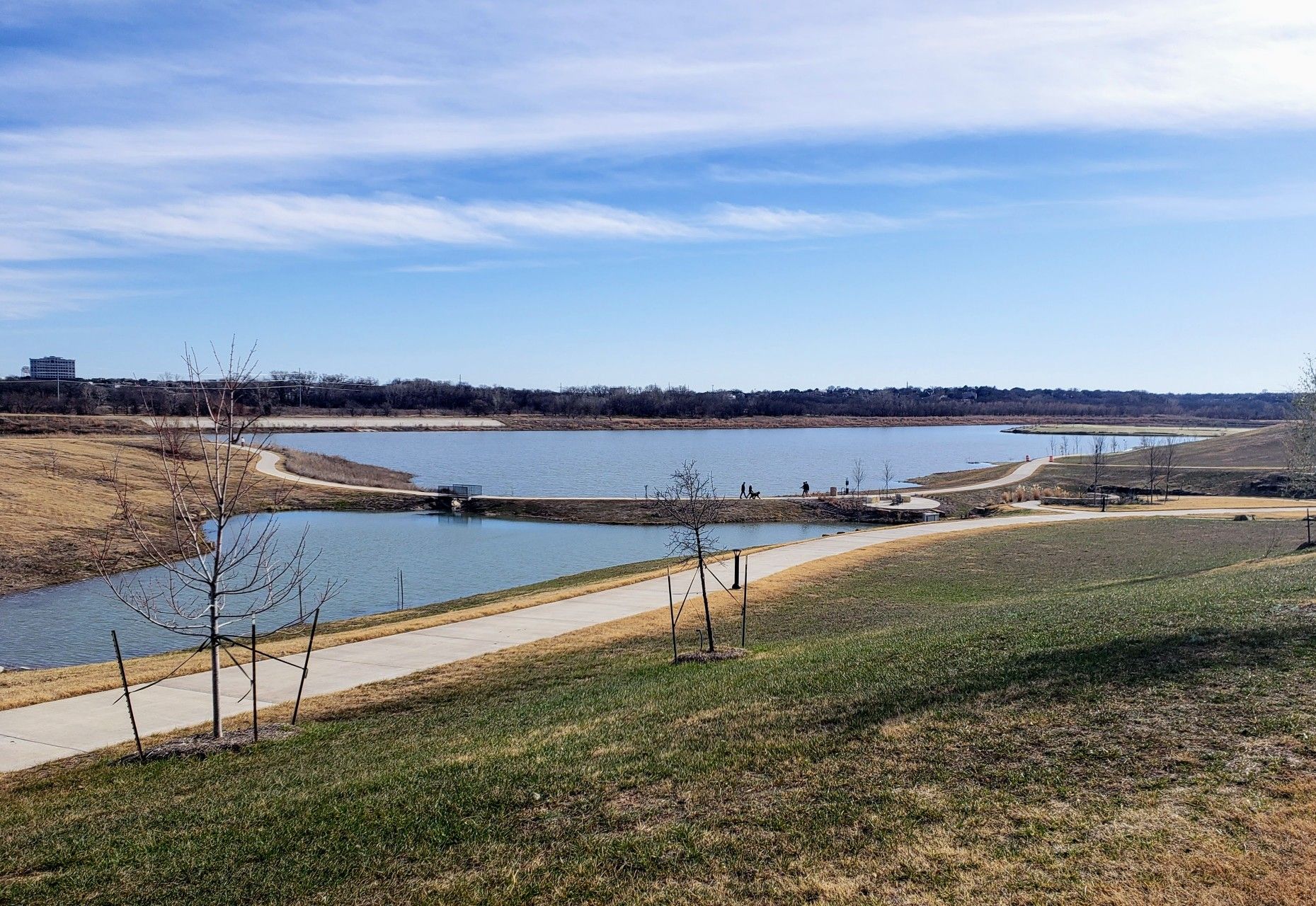 Wide view of a lake with walking paths and distant people under a blue sky. - River Legacy Trail