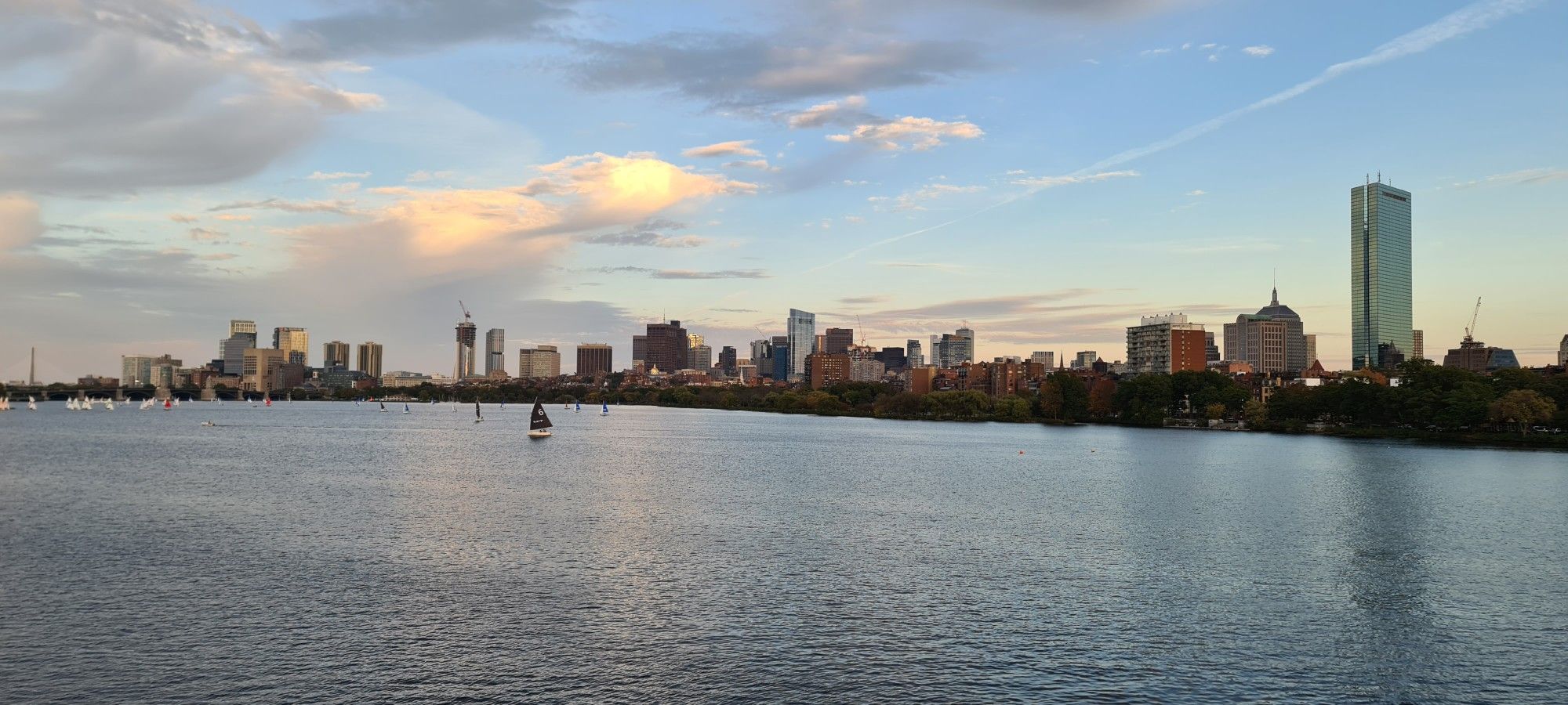 Golden hour light illuminates a city skyline across a river with a sailboat. - Lexington Battle Green – Spy Pond loop from Hynes Convention Center
