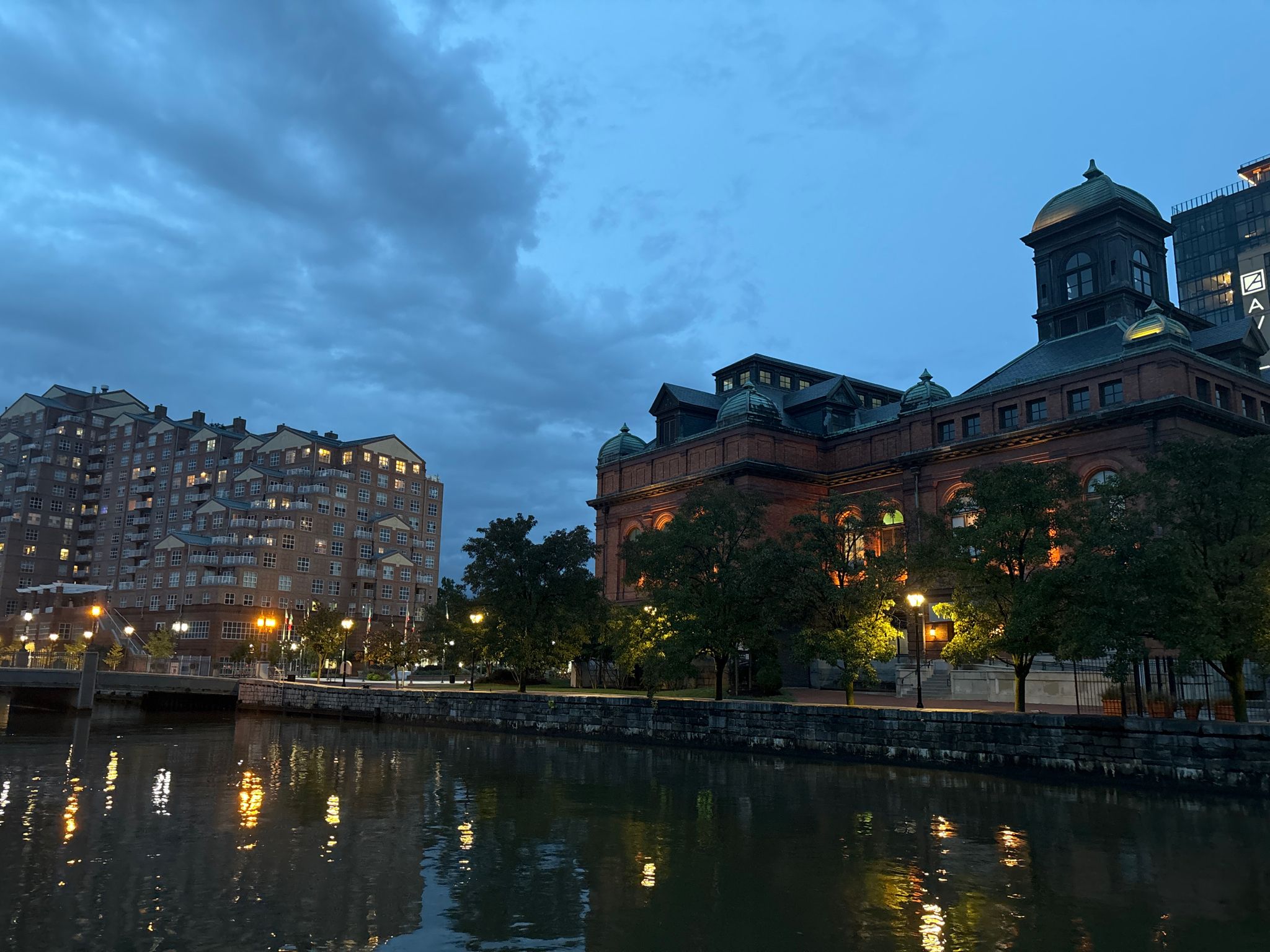 Historic brick building and apartment complex by water at dusk, with lights reflecting. - Seven Foot Knoll Lighthouse – Jones Falls loop from Highlandtown