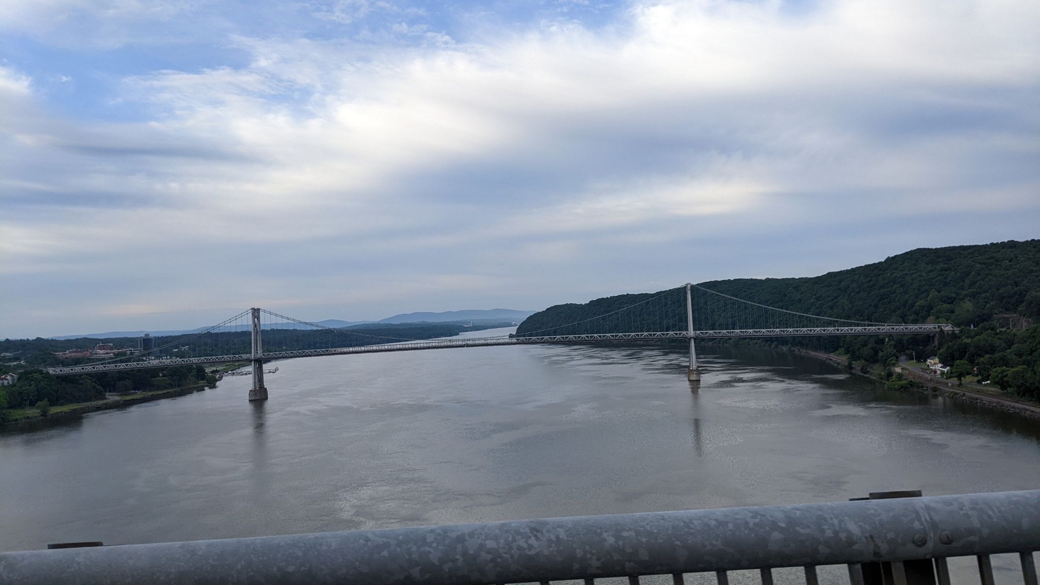 A suspension bridge spans a wide river with forested hills under a cloudy sky. - Hudson Valley Rail Trail and Walkway Over the Hudson Loop
