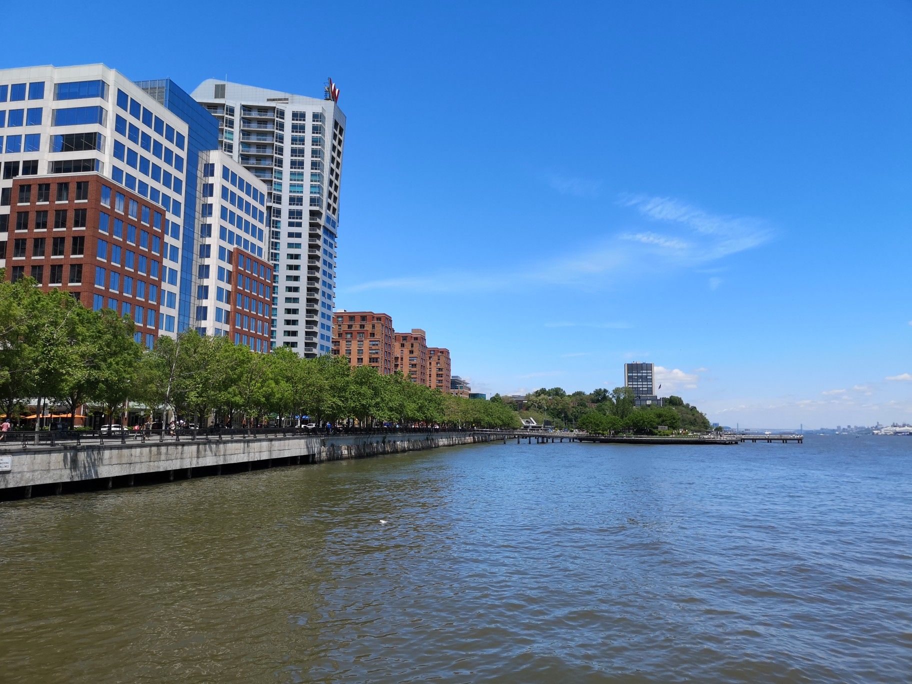 Waterfront promenade with trees and tall buildings under a clear blue sky. - Hoboken Terminal – Weehawken Cove loop from 2nd Street