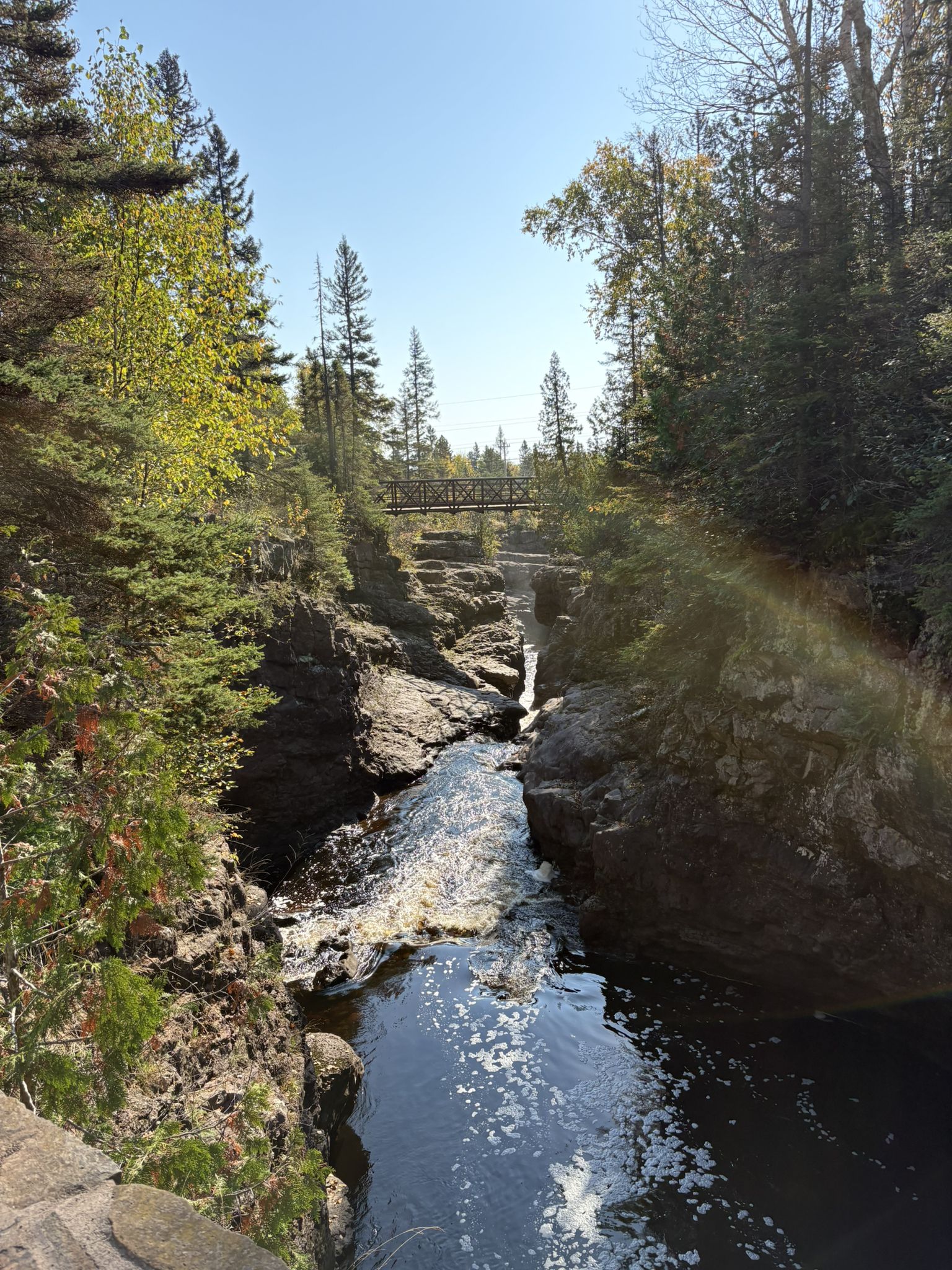 A river flows through a rocky gorge with a bridge in the distance, sunny. - Temperance River Falls Loop