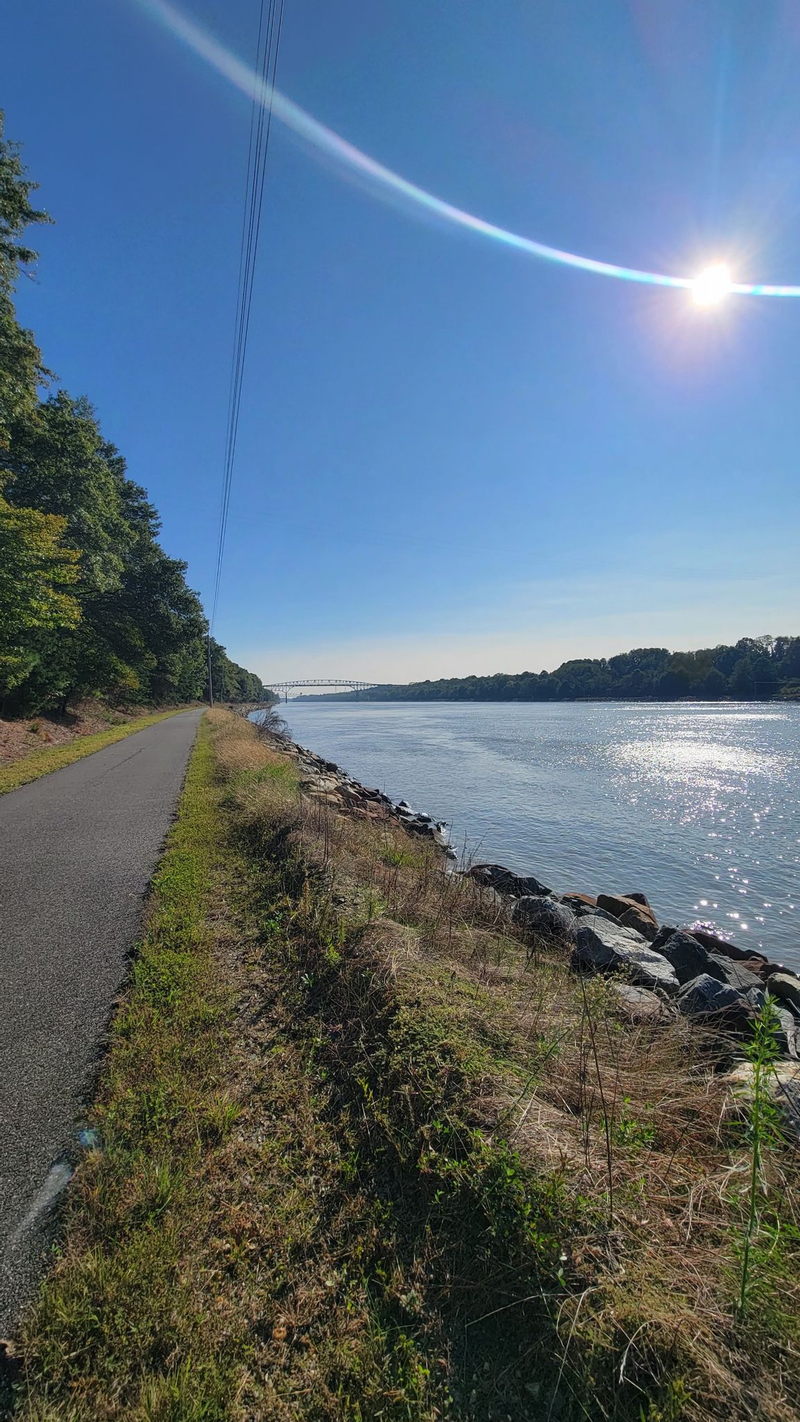A sunny river view from a paved path with bright sun and blue sky. - Running loop from Summit Bridge