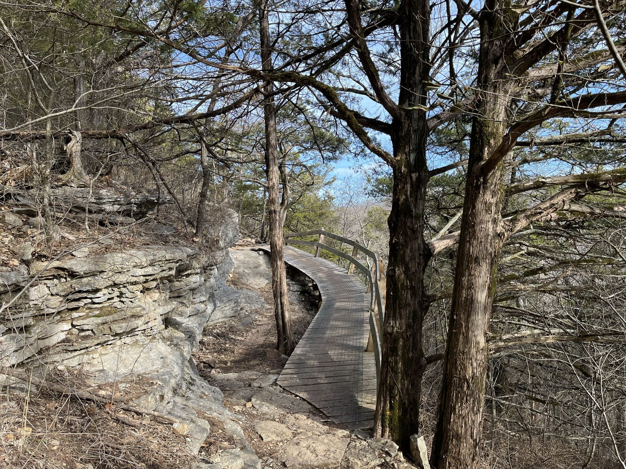 A wooden boardwalk curves through a rocky, sunlit forest with bare trees. - Blowing Springs and Wonderland Loop