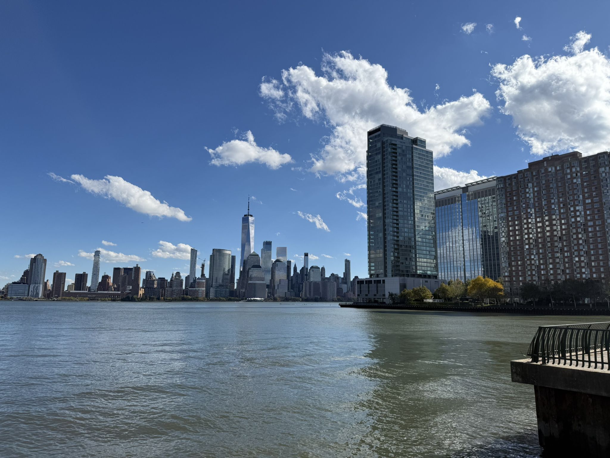 City skyline across water with foreground buildings under a sunny, blue sky. - LeFrak Point Lighthouse – Hoboken Newport Walkway loop from Grove Street