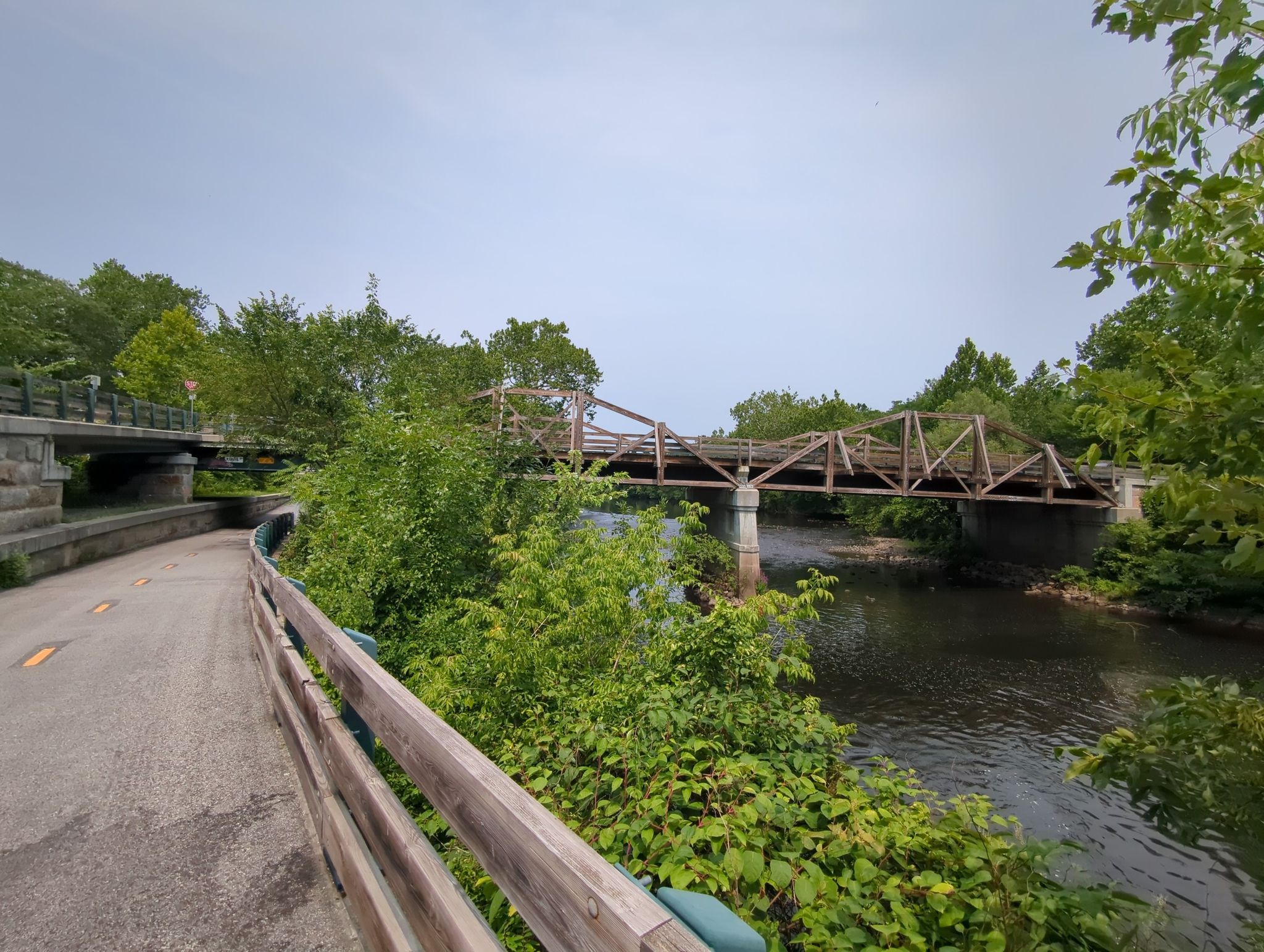 A paved path curves beside a river with two wooden bridges ahead. - Running loop from Valley Falls
