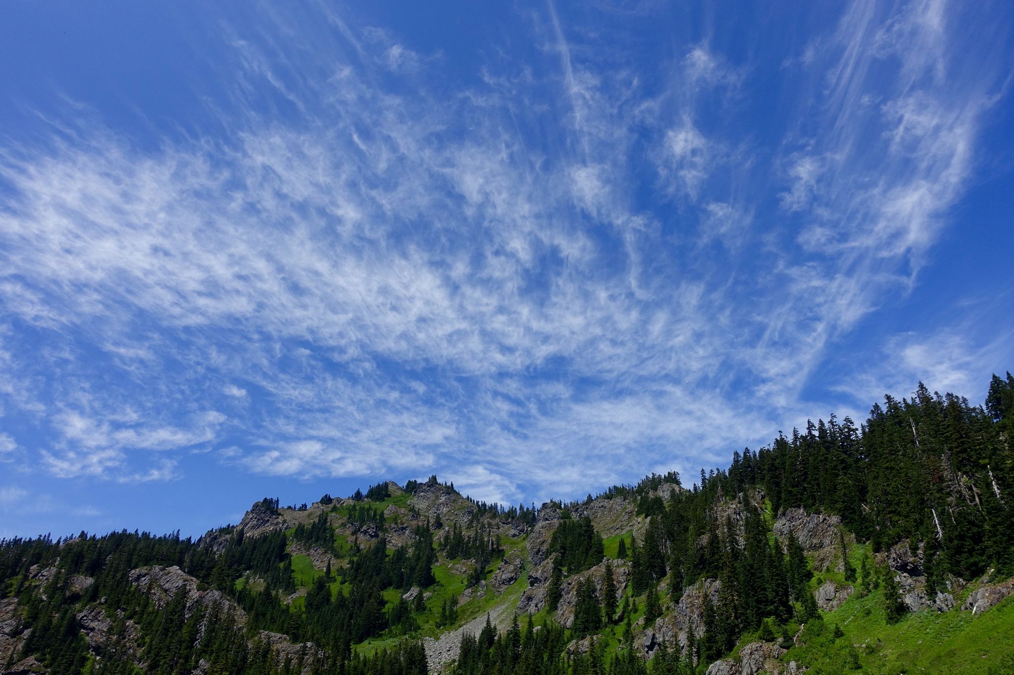 Bright blue sky with wispy clouds above a rocky, forested mountain ridge. - Rampart Lakes and Alta Mountain Trail