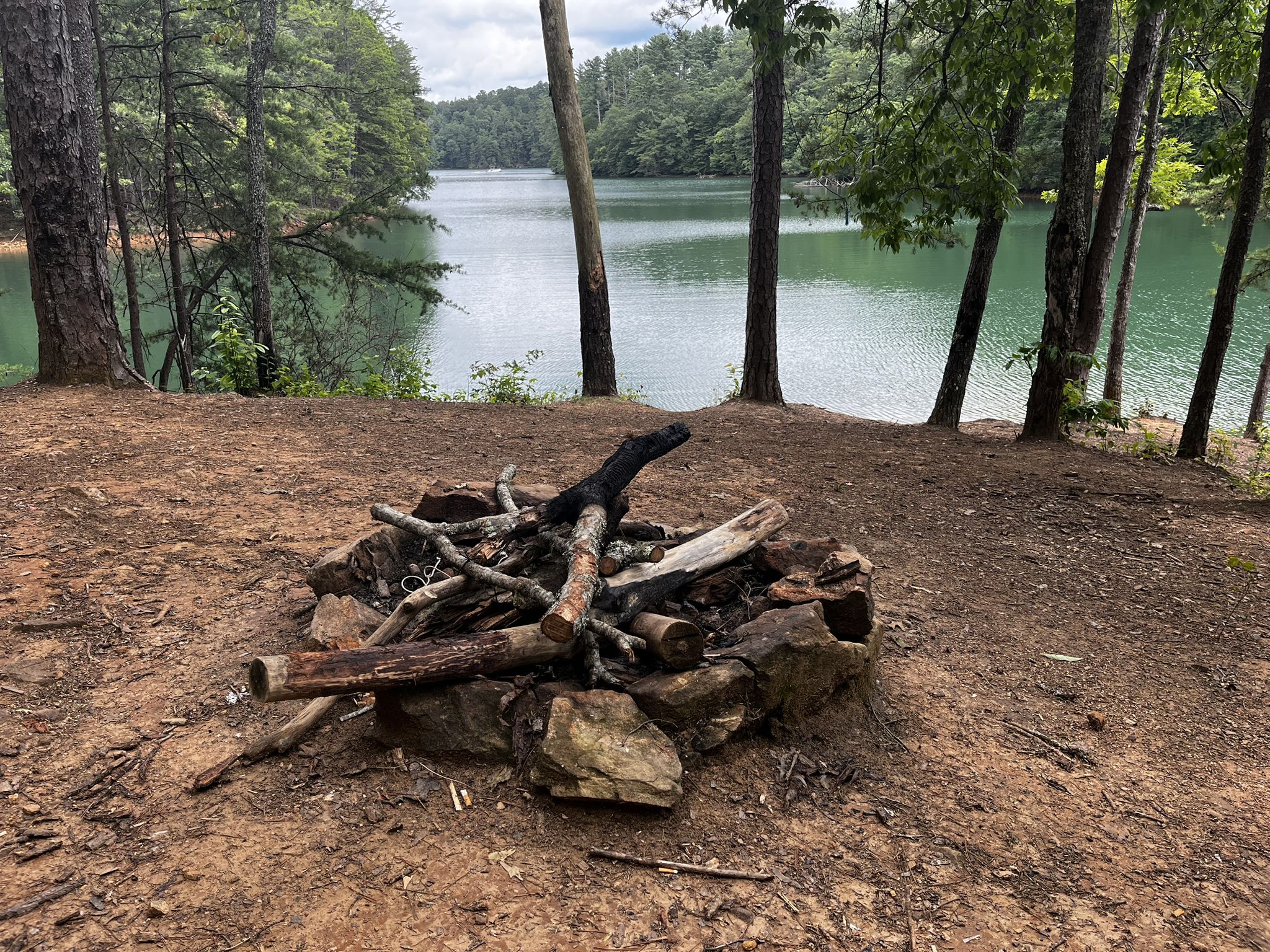A rock fire pit with wood, beside a lake surrounded by green trees. - Green Mountain and Lake Blue Ridge Loop
