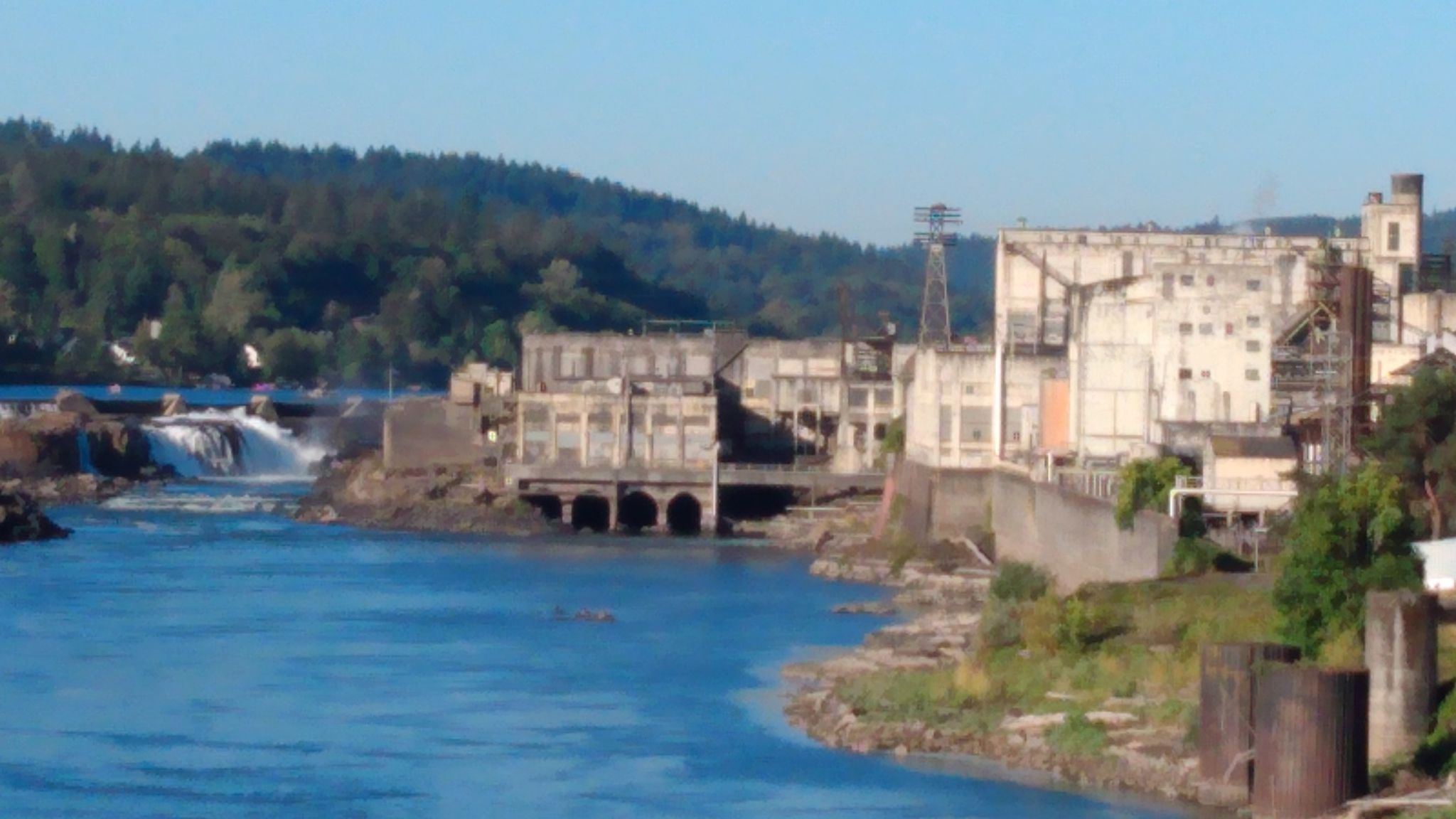 River with waterfalls next to large industrial buildings and forested hills. - Logging County Road – McLoughlin House Site loop from River Bluff Open Space