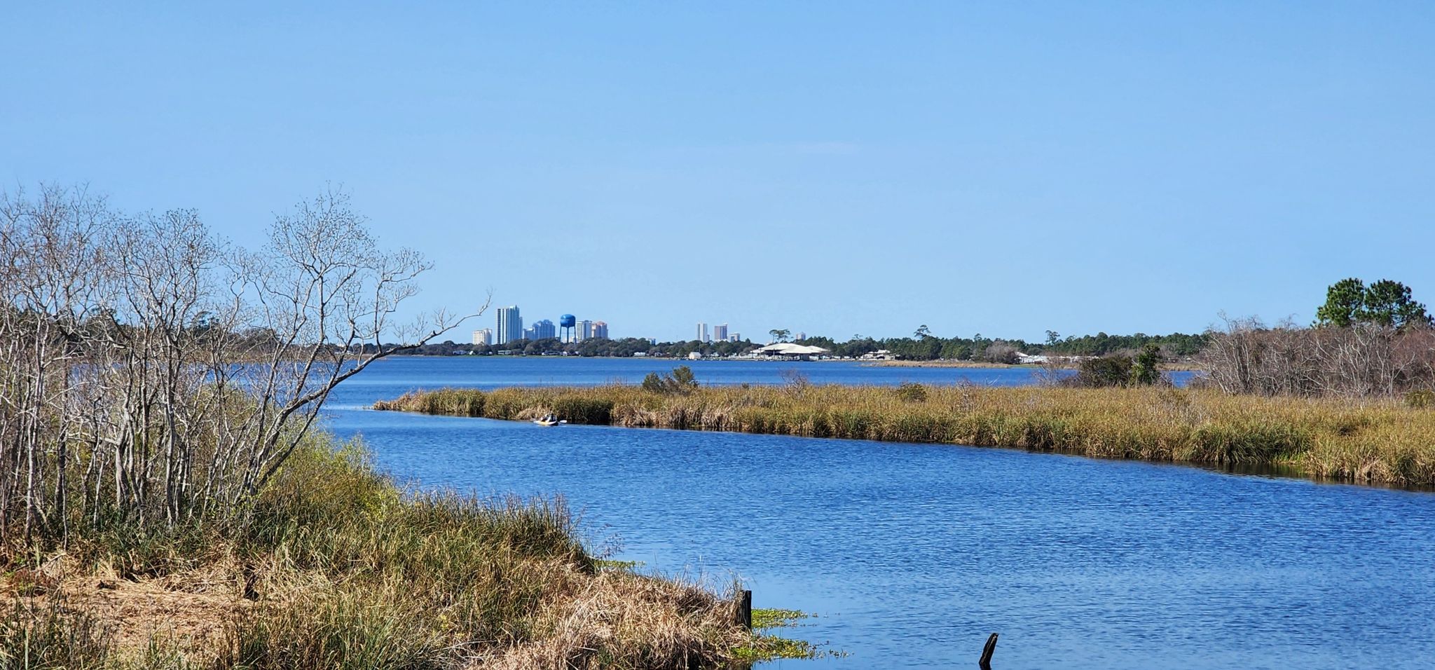 A wide river with grassy banks leads to a distant city skyline. - Middle Lake – Middle River loop from Gulf State Park