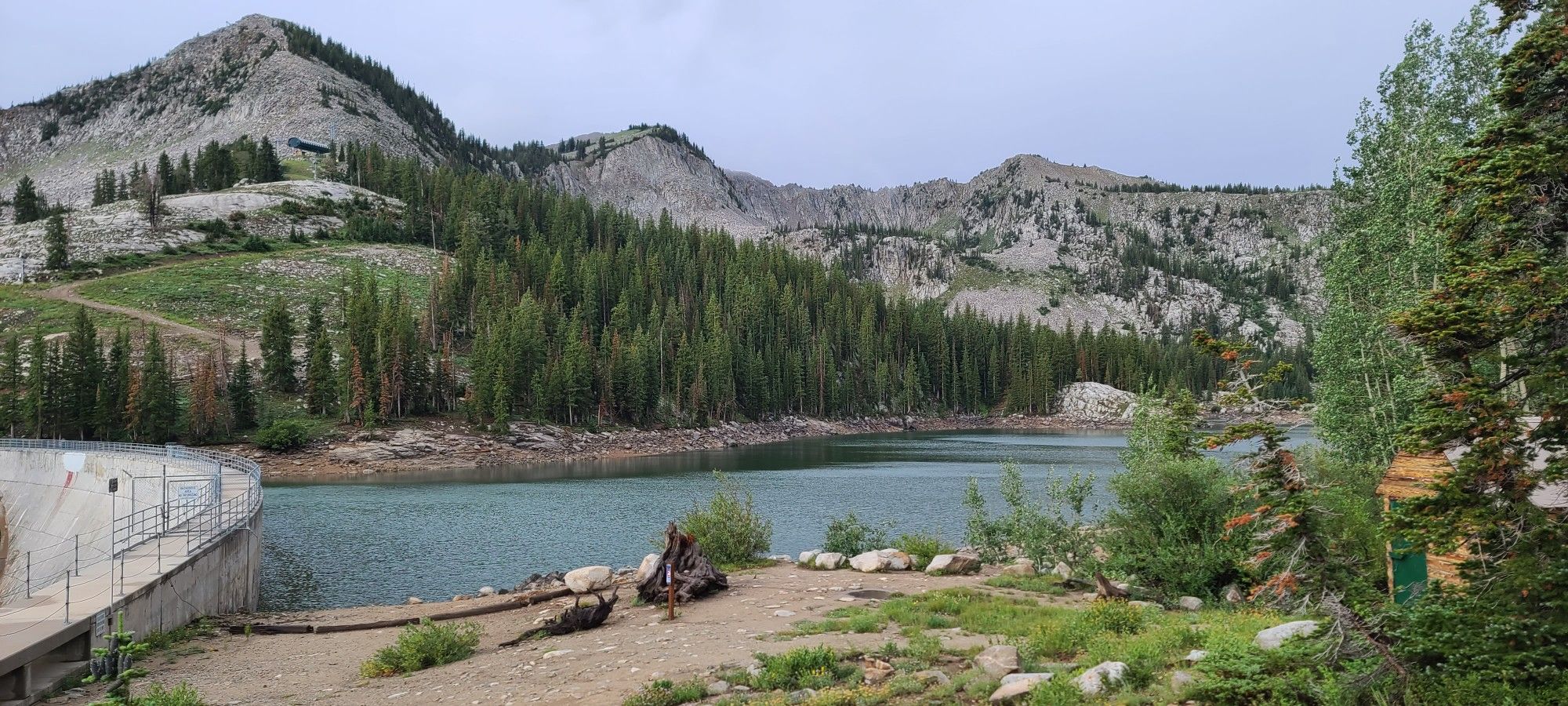 A mountain lake with a dam, surrounded by forests and rocky peaks under a cloudy sky. - Alta-Brighton Lakes Loop