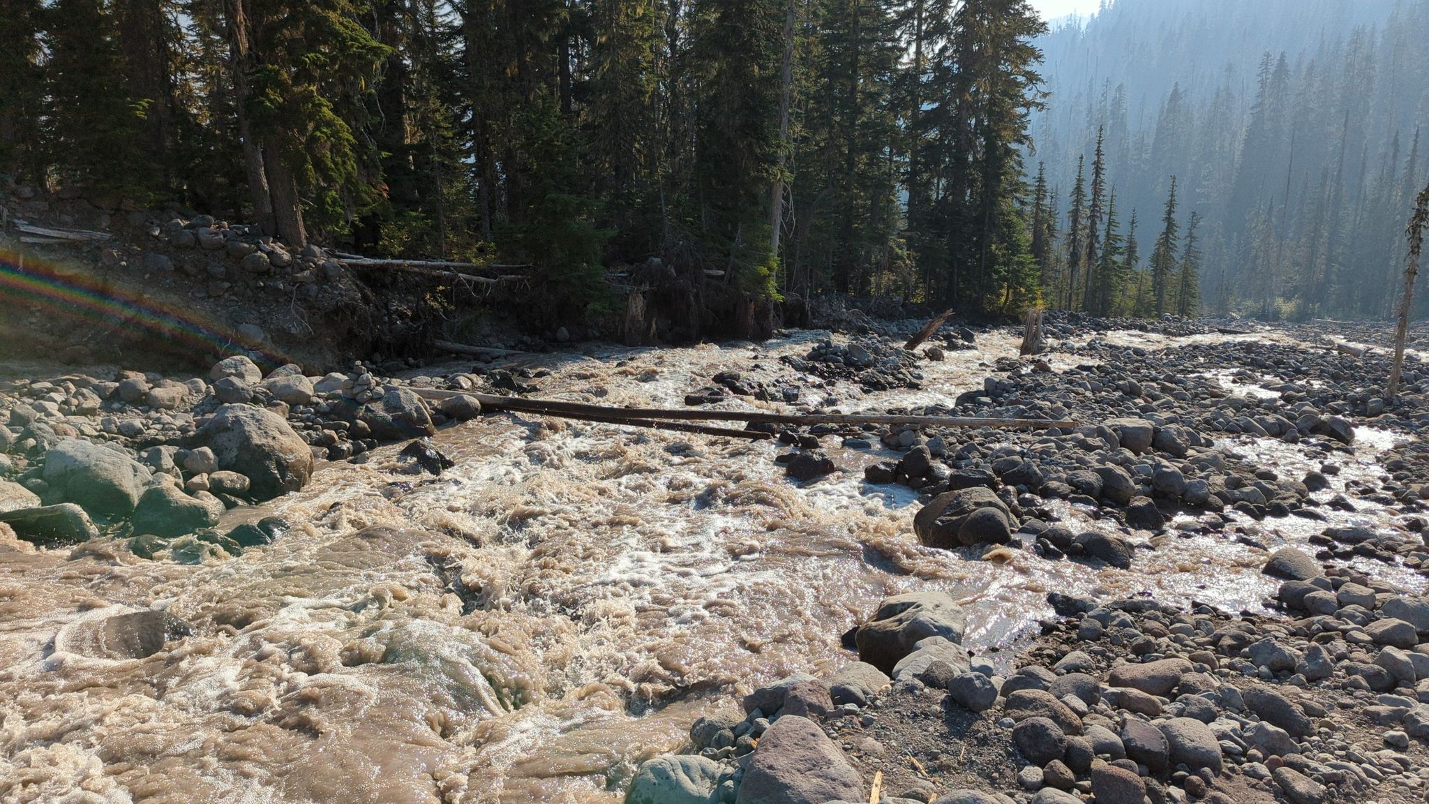 Wide, muddy river flowing over rocks through a dense forest on a hazy day. - Park Butte Trail