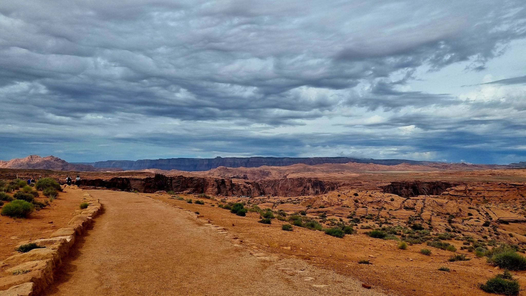 Dirt path leading towards a distant canyon under a cloudy sky with people. - Horseshoe Bend Trailhead – View of Horseshoe Bend loop from Horseshoe Bend